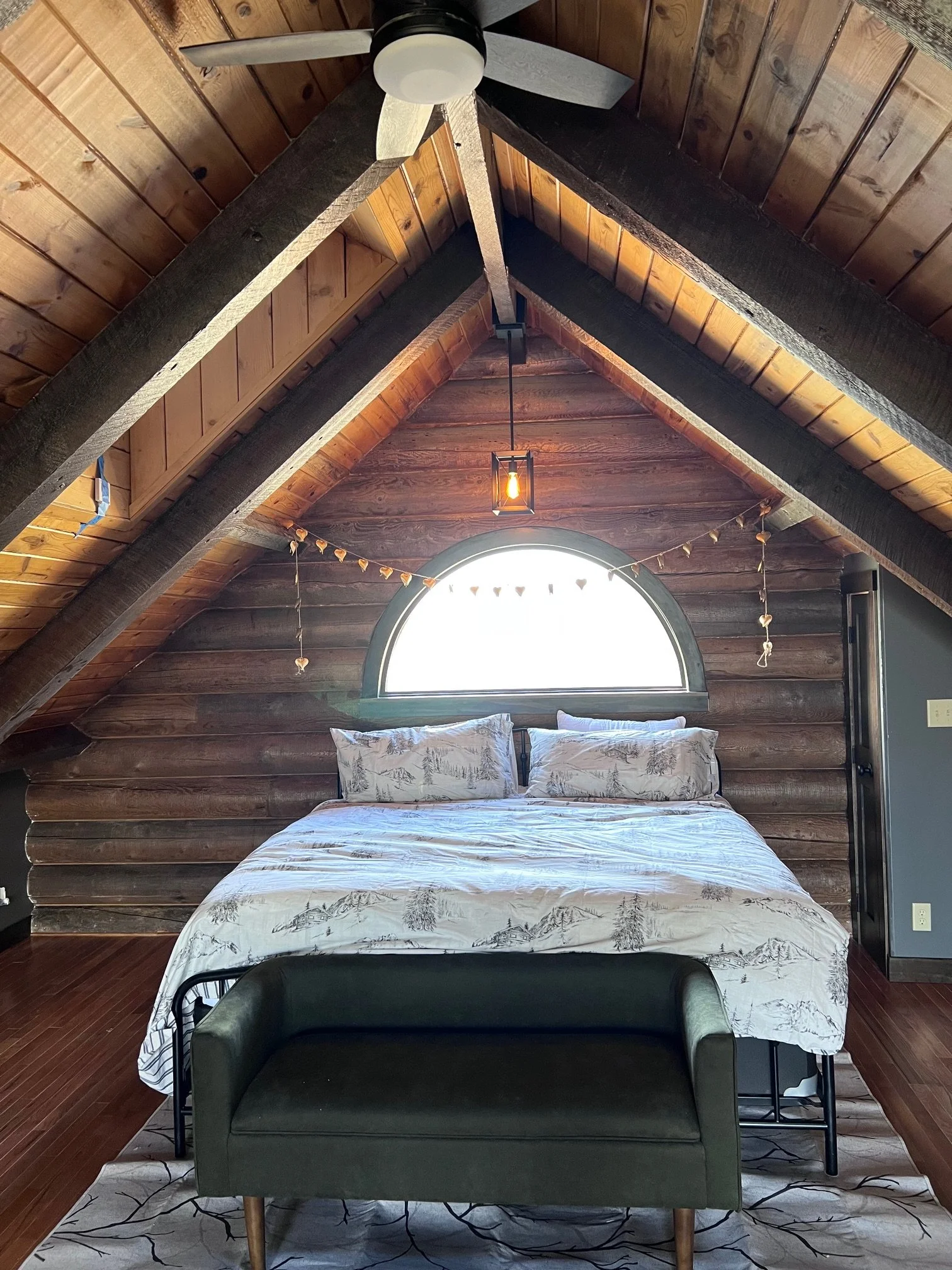 A bedroom with vaulted wooden ceilings and wall paneling, a window with an arched top, a bed with mountain and tree-patterned bedding, a gray sofa at the foot of the bed, and a ceiling fan with a light fixture.
