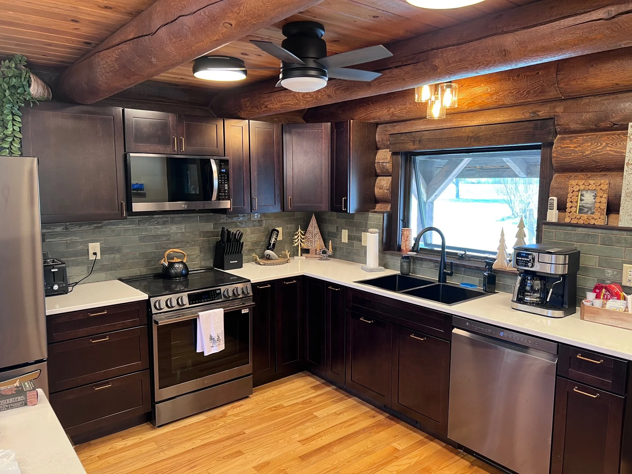 A cozy kitchen with wooden ceiling beams and walls. Dark wooden cabinets, stainless steel appliances including a microwave and stove, and a countertop with small decorative trees. Large window with view of snow outside. Ceiling fan and light fixtures add to the warm ambiance.