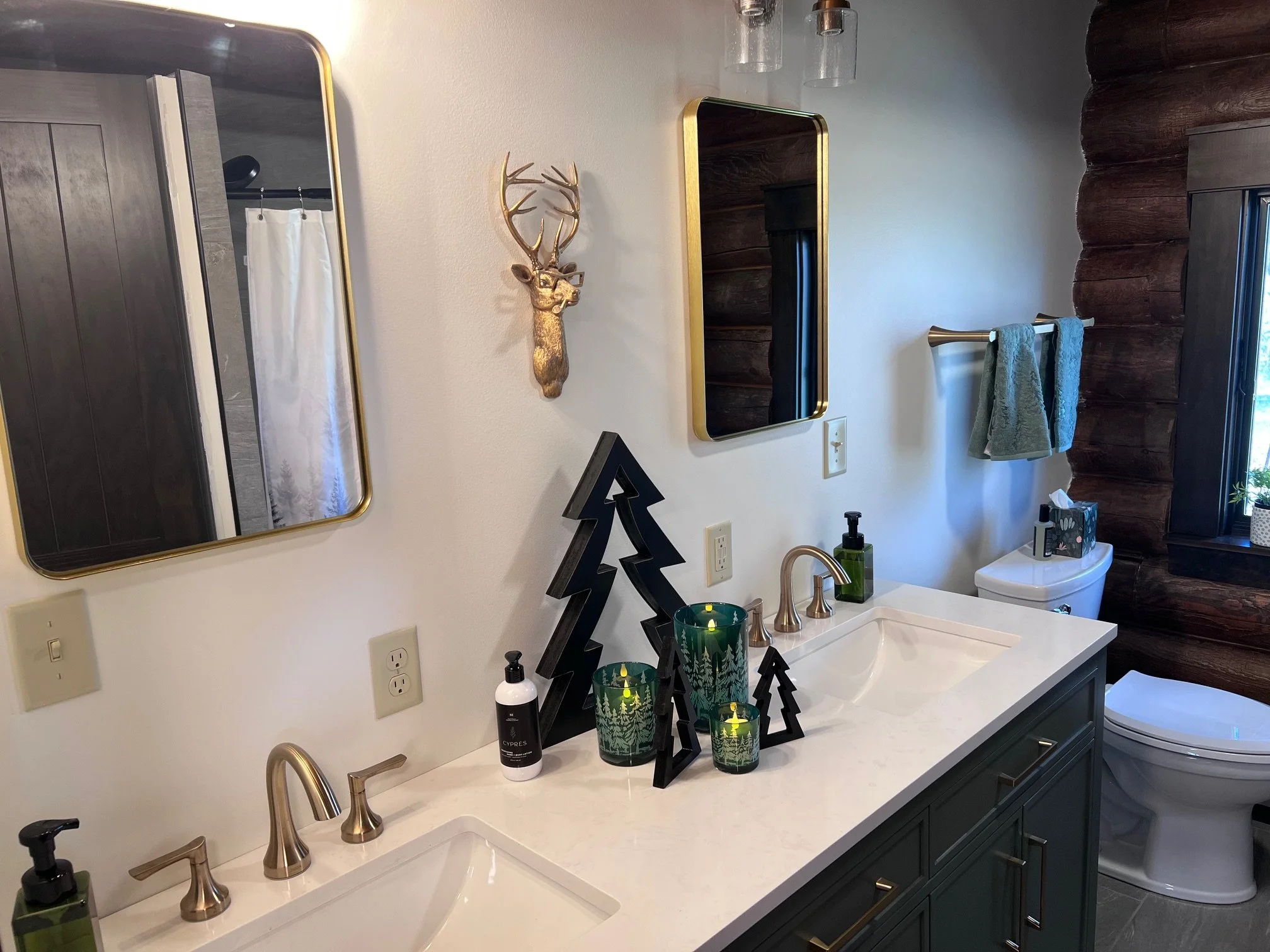 Bathroom vanity with two sinks, two mirrors with gold frames, wall-mounted deer head decoration, black tree-shaped decor, candles, soap dispenser, and towels, with a toilet and window in the background.