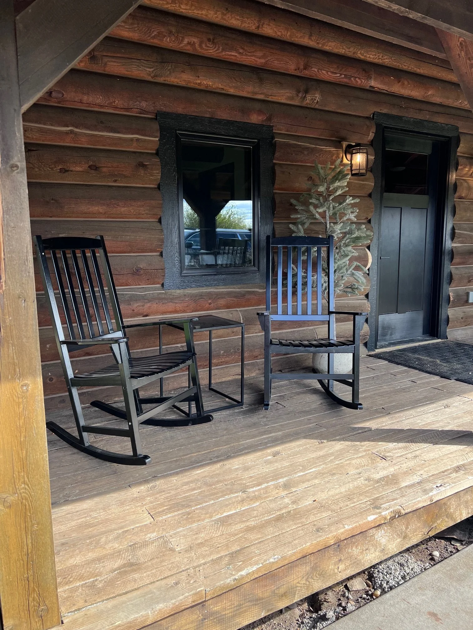 A porch area with two black rocking chairs and a small black table, a small potted evergreen tree, a black window and a black door with a lantern-style light fixture, all against a log cabin wall.