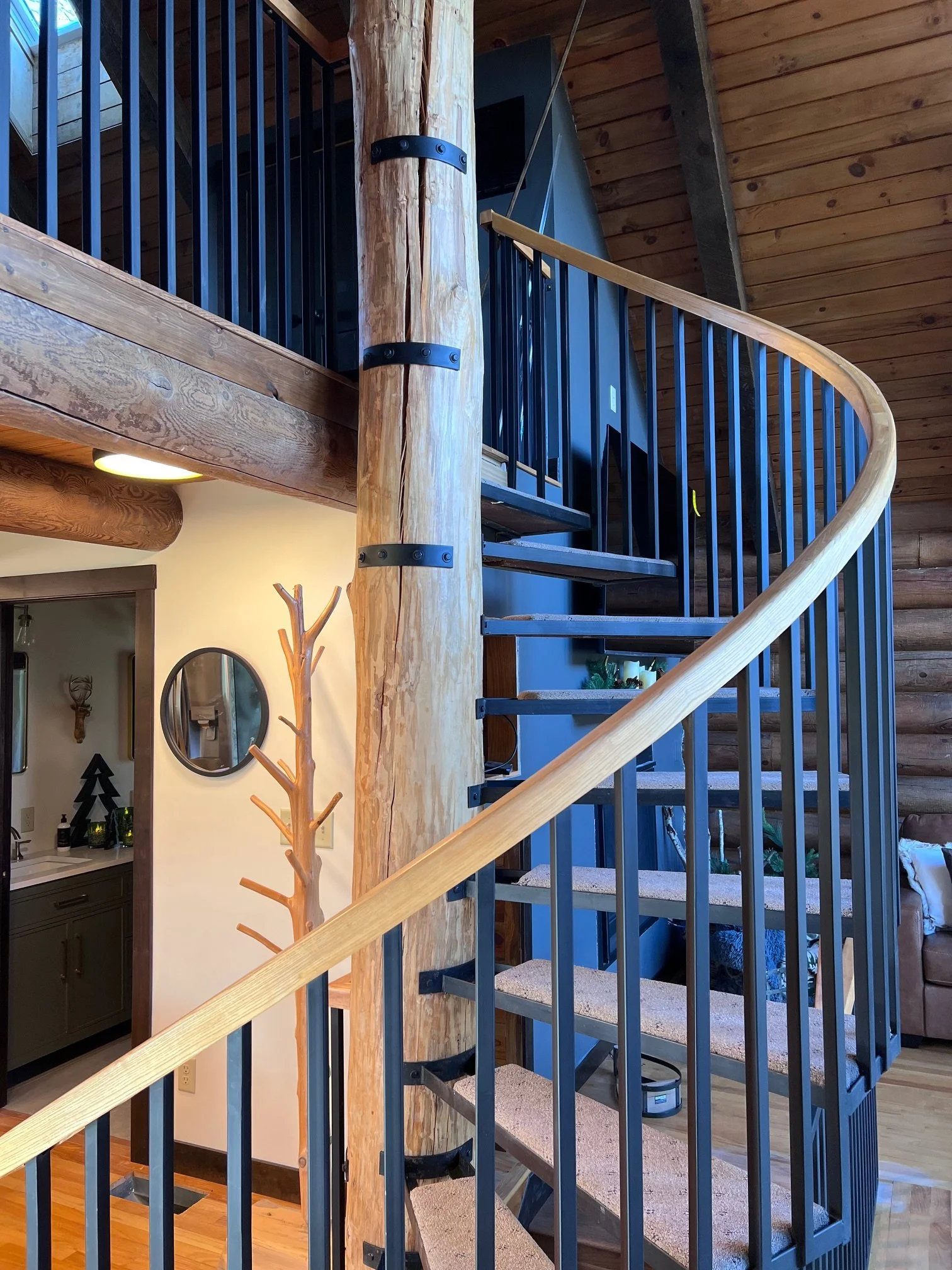 Interior view of a staircase in a house with wooden walls and ceiling, featuring black metal railings and a natural wood handrail, with a nearby room visible through the doorway with a coat rack and wall decor.