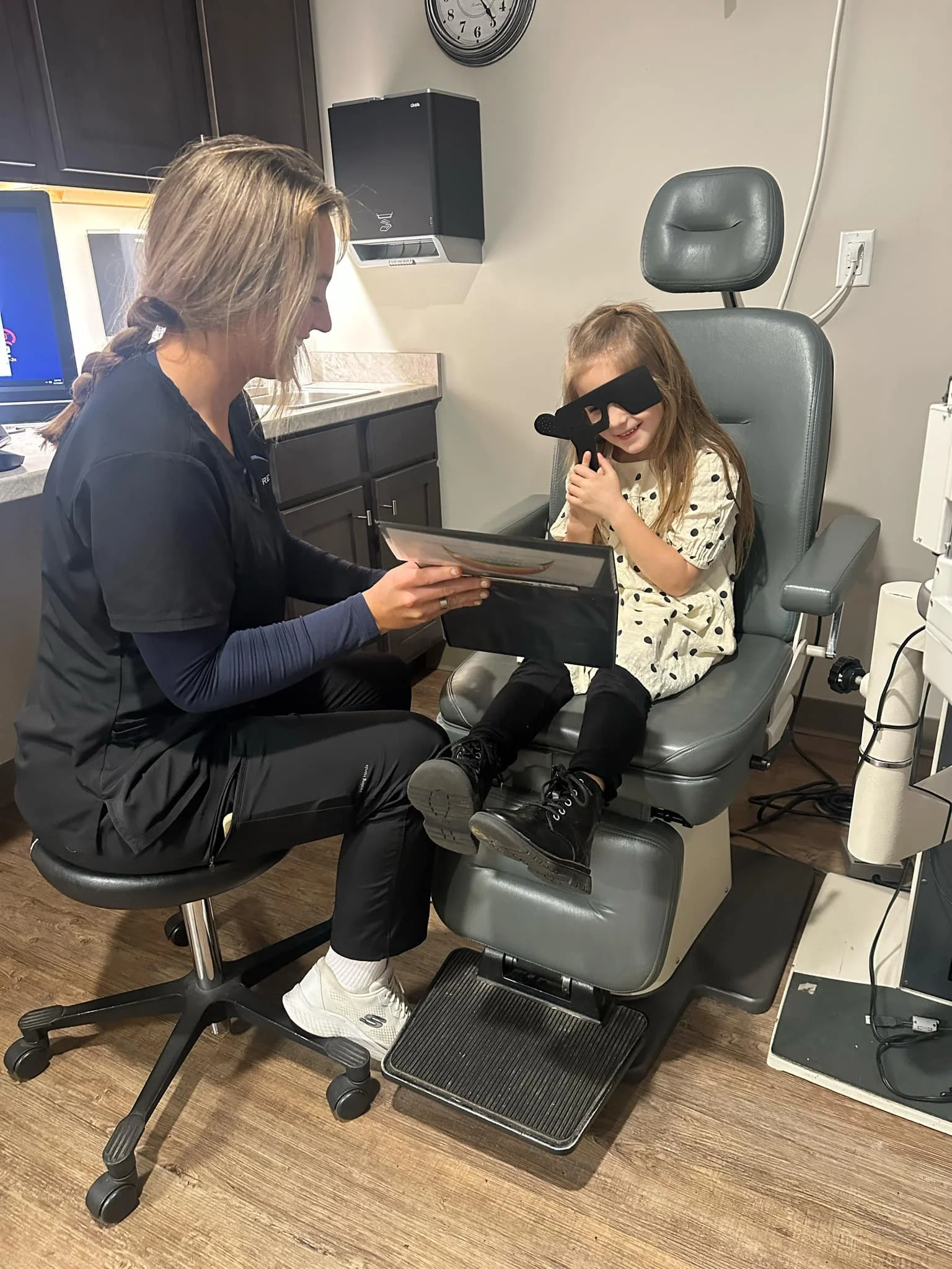 A child sitting in an optometrist's chair during an eye exam while an adult sits nearby holding a chart.