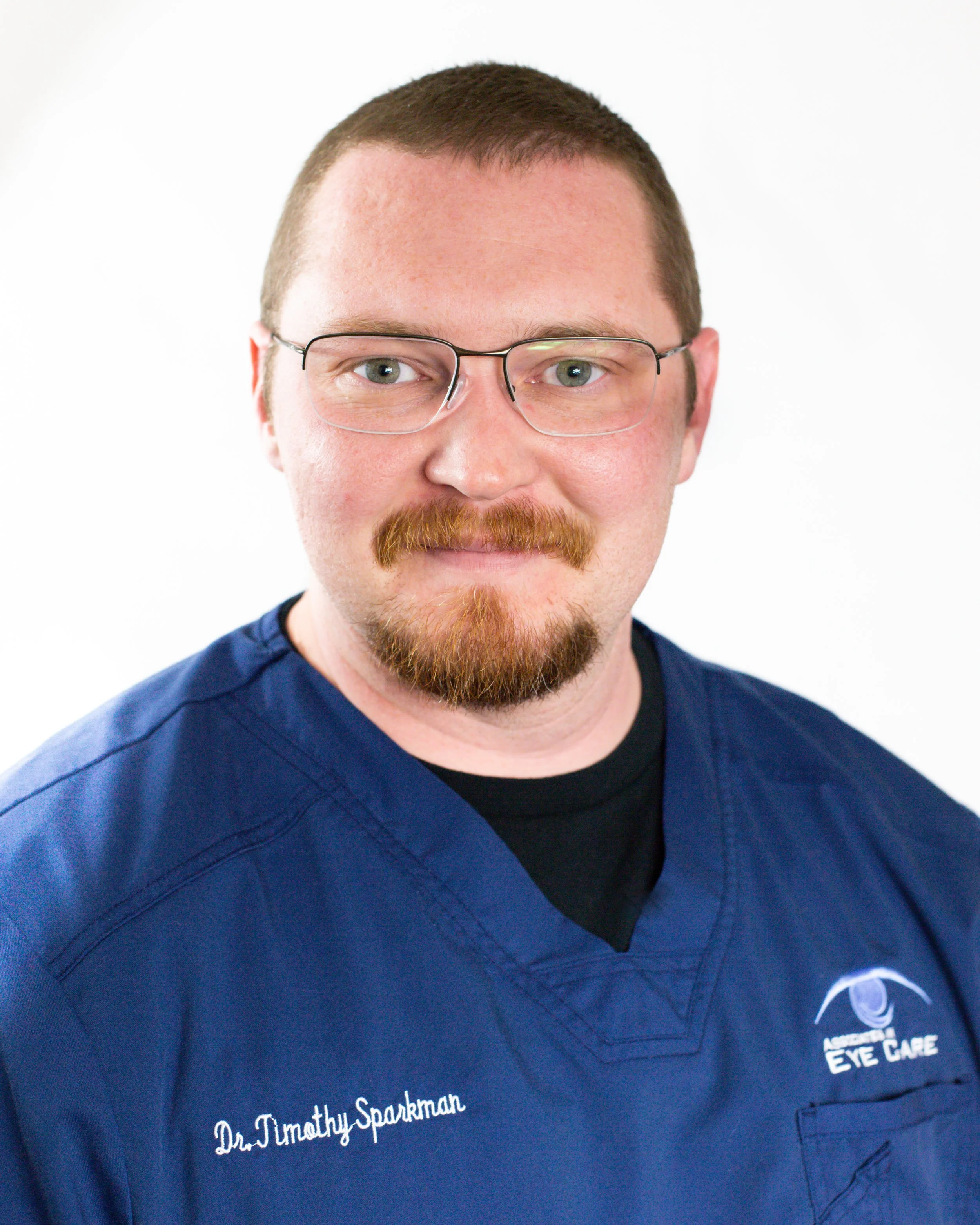 A man wearing a blue medical scrub with glasses and a beard, standing against a plain white background.