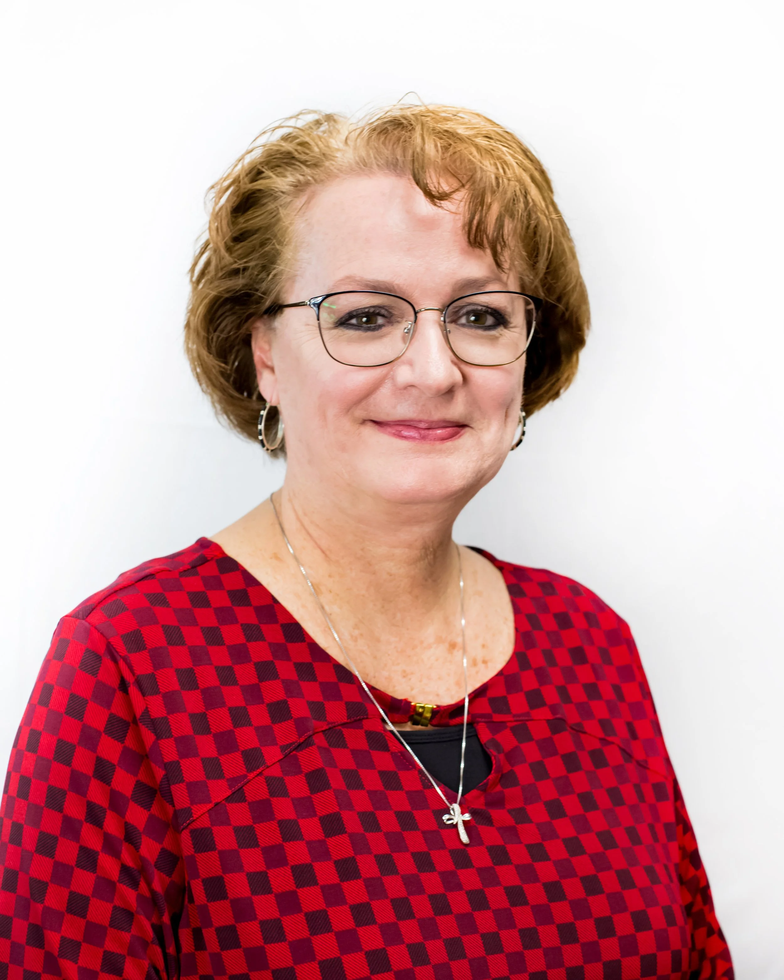 Woman wearing glasses and a red checkered top, smiling, with short reddish-brown hair, against a white background.