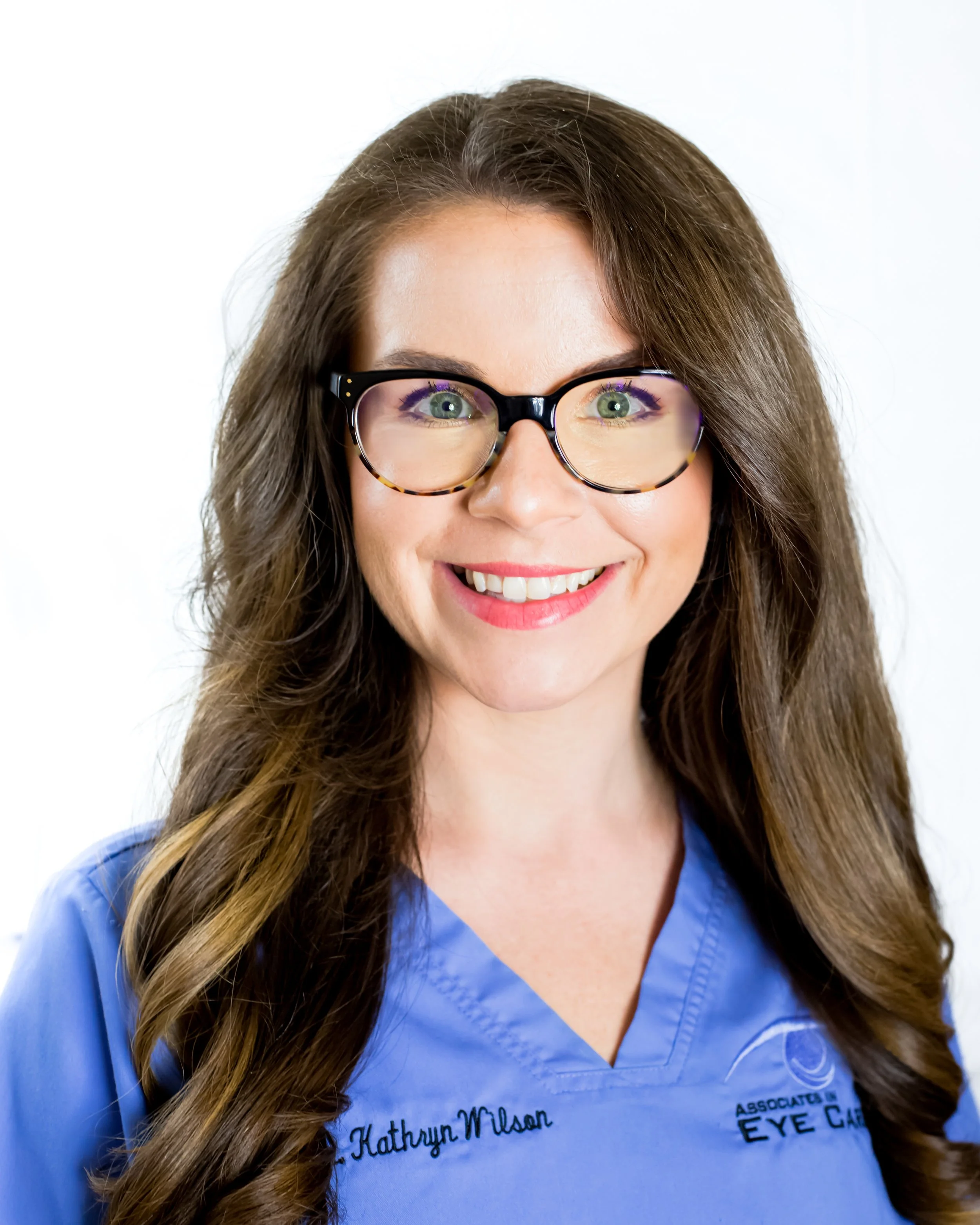 A smiling woman with long brown hair wearing glasses and a blue scrub top embroidered with eye care details.
