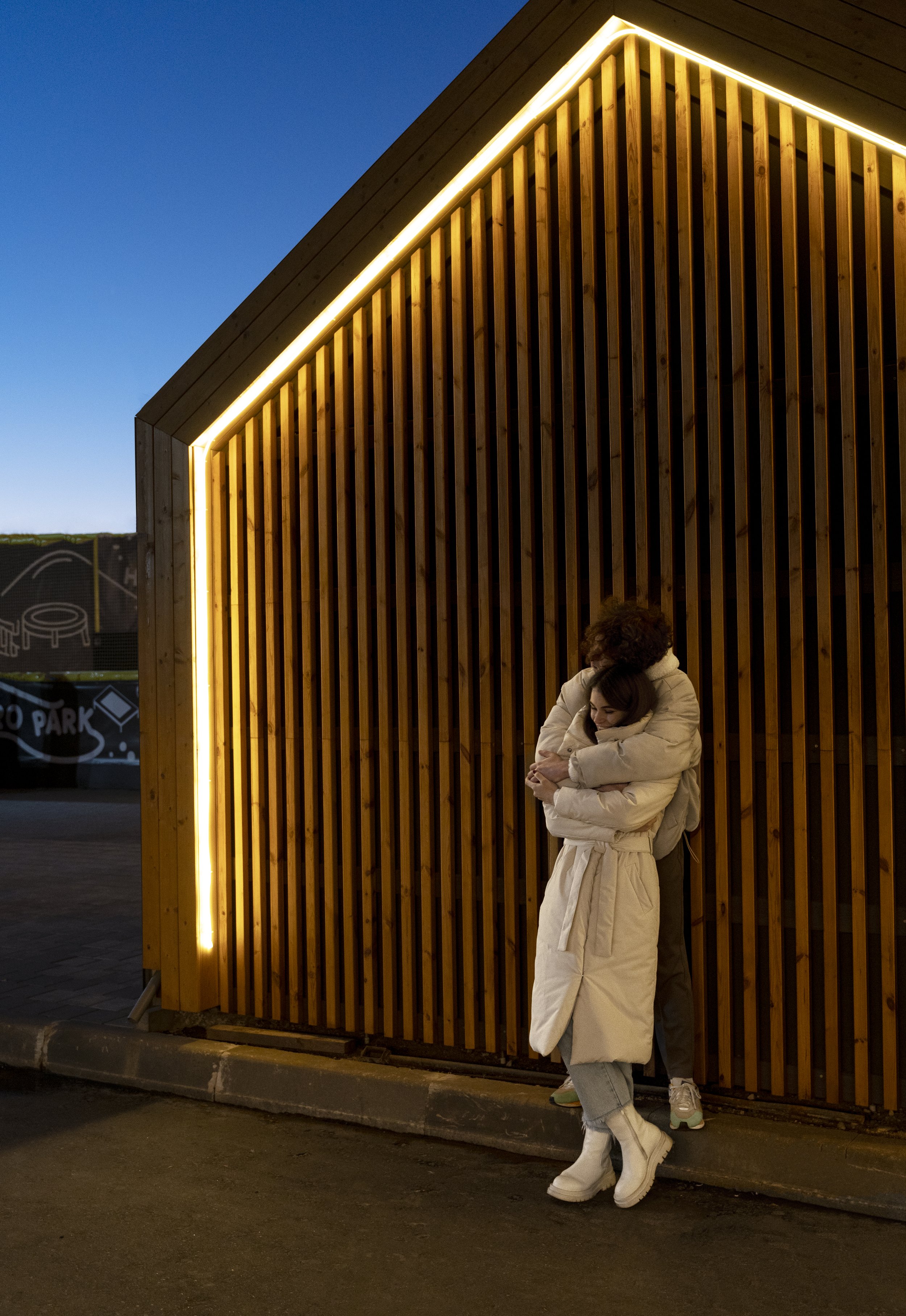 Two people hugging near a wooden wall with lighting, outdoor setting, evening or night.