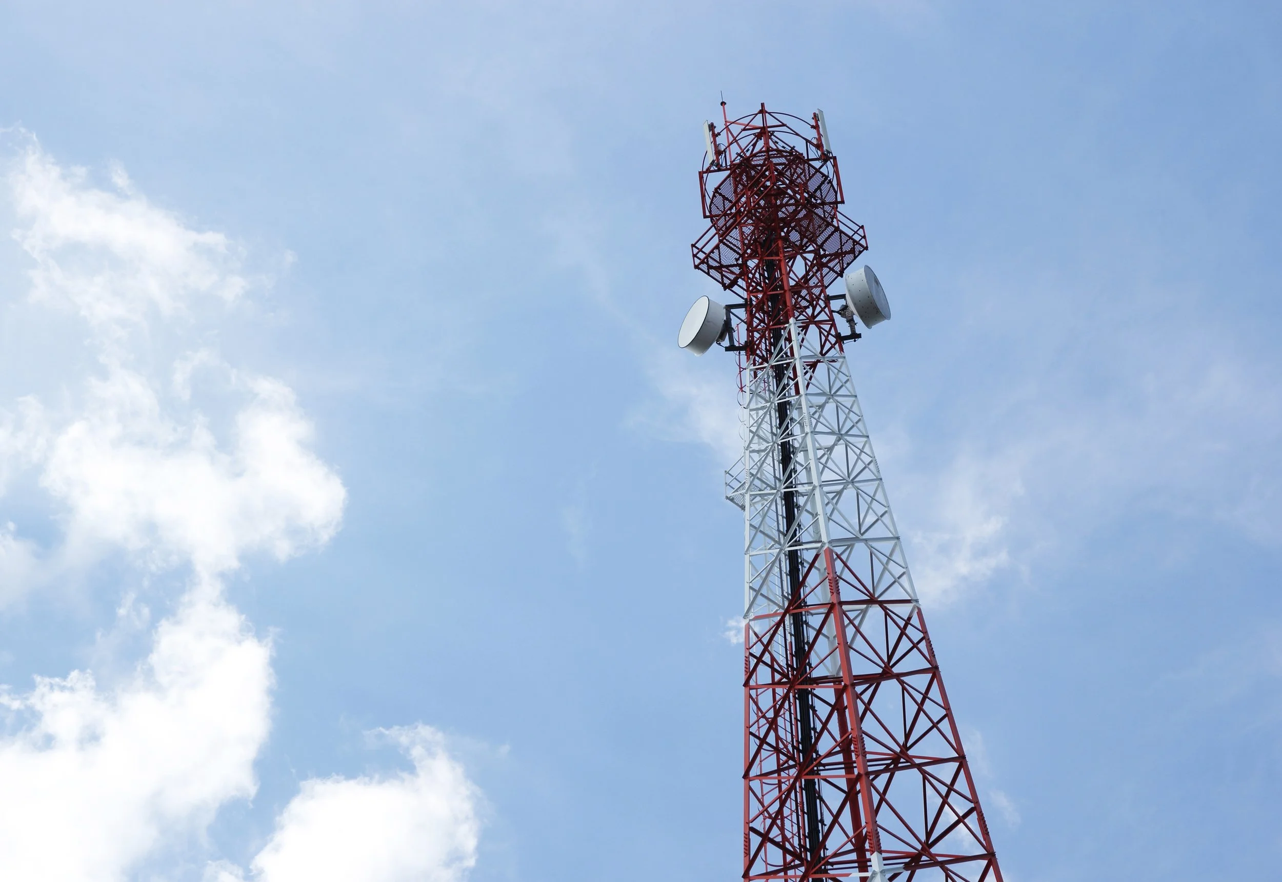 A worker on a green lift works on a telecommunications tower with multiple antennas and equipment against a pale sky.