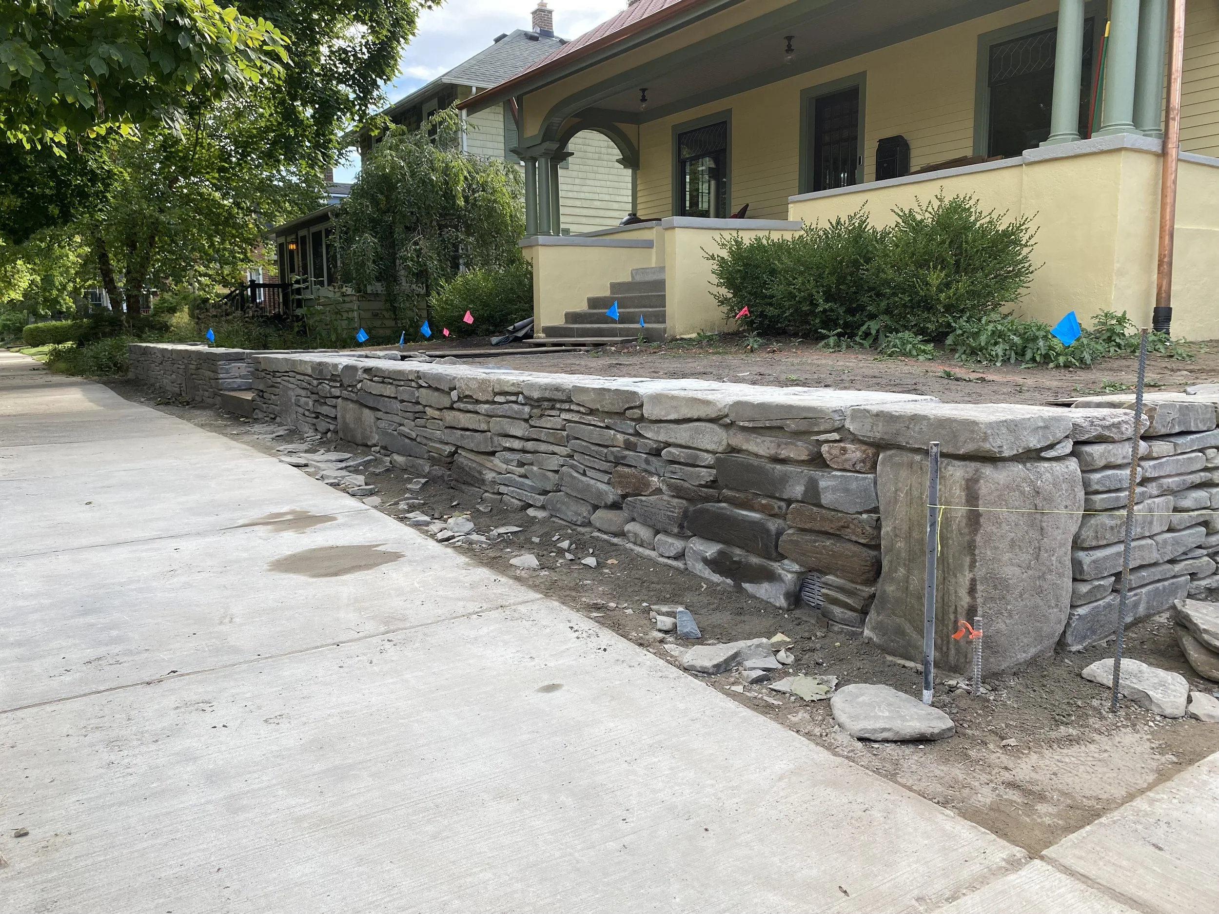 Front corner stone retaining wall mid-construction, courses of fieldstone set by hand where the concrete curb once stood