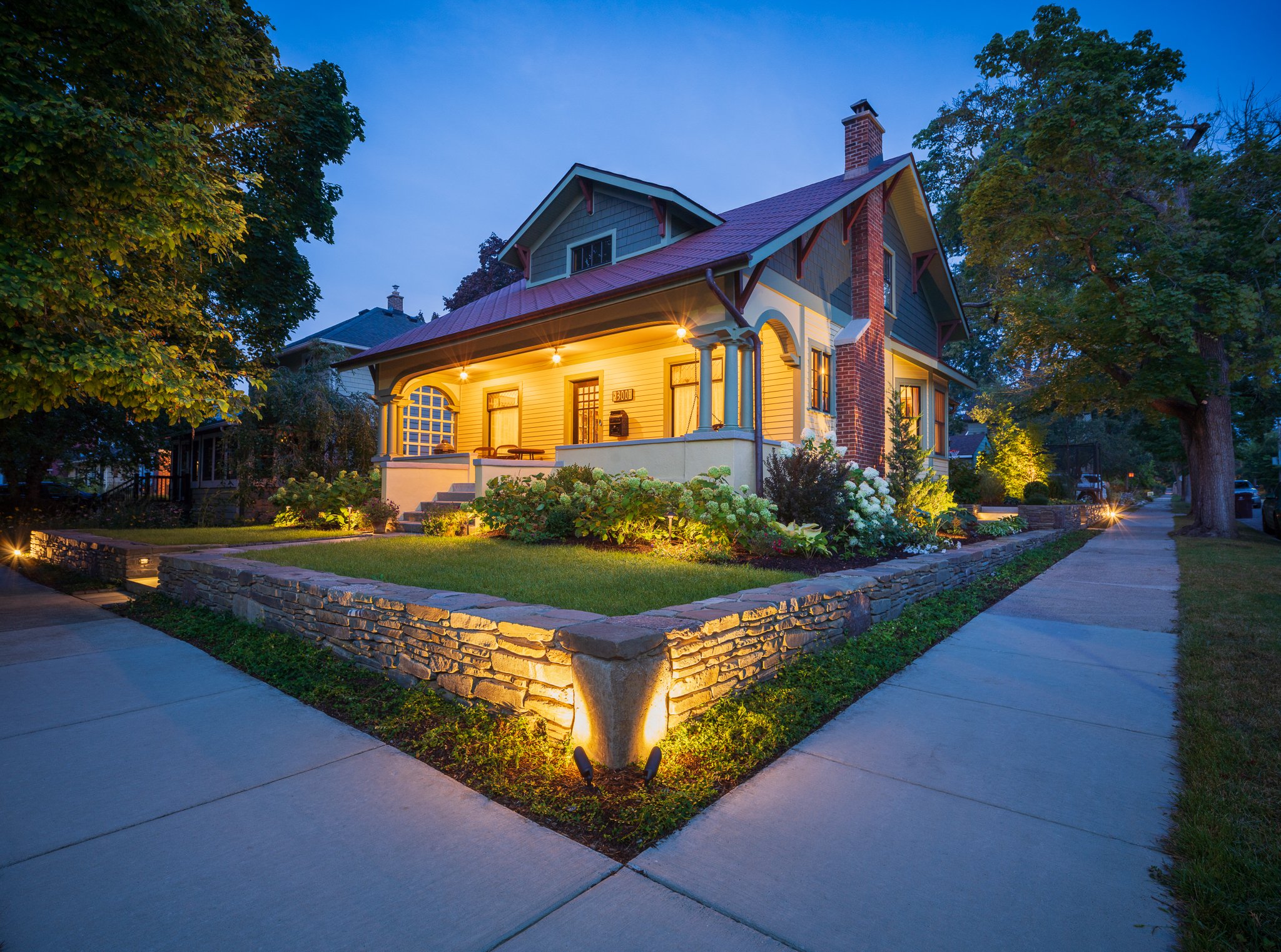 The finished front corner at dusk — an uplit dry-stacked fieldstone retaining wall wrapping layered garden beds below the restored Arts and Crafts home