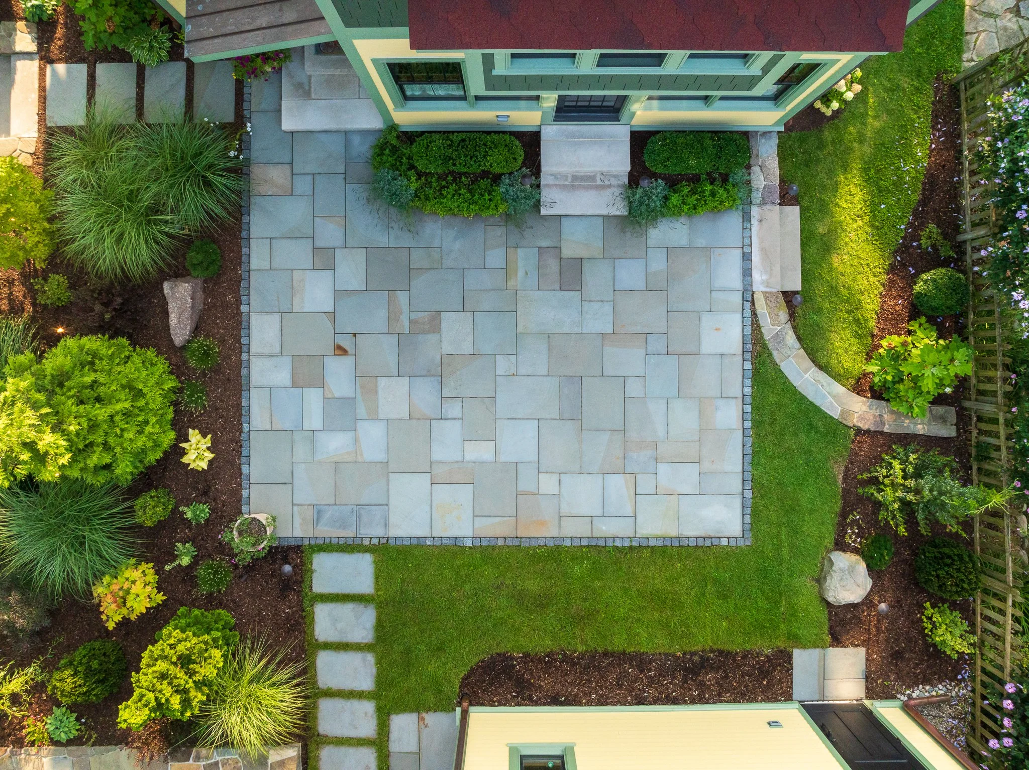 A backyard patio with a stone tile surface surrounded by green bushes, plants, and a small section of grass, with a house painted yellow with green trim and a red roof in the background.