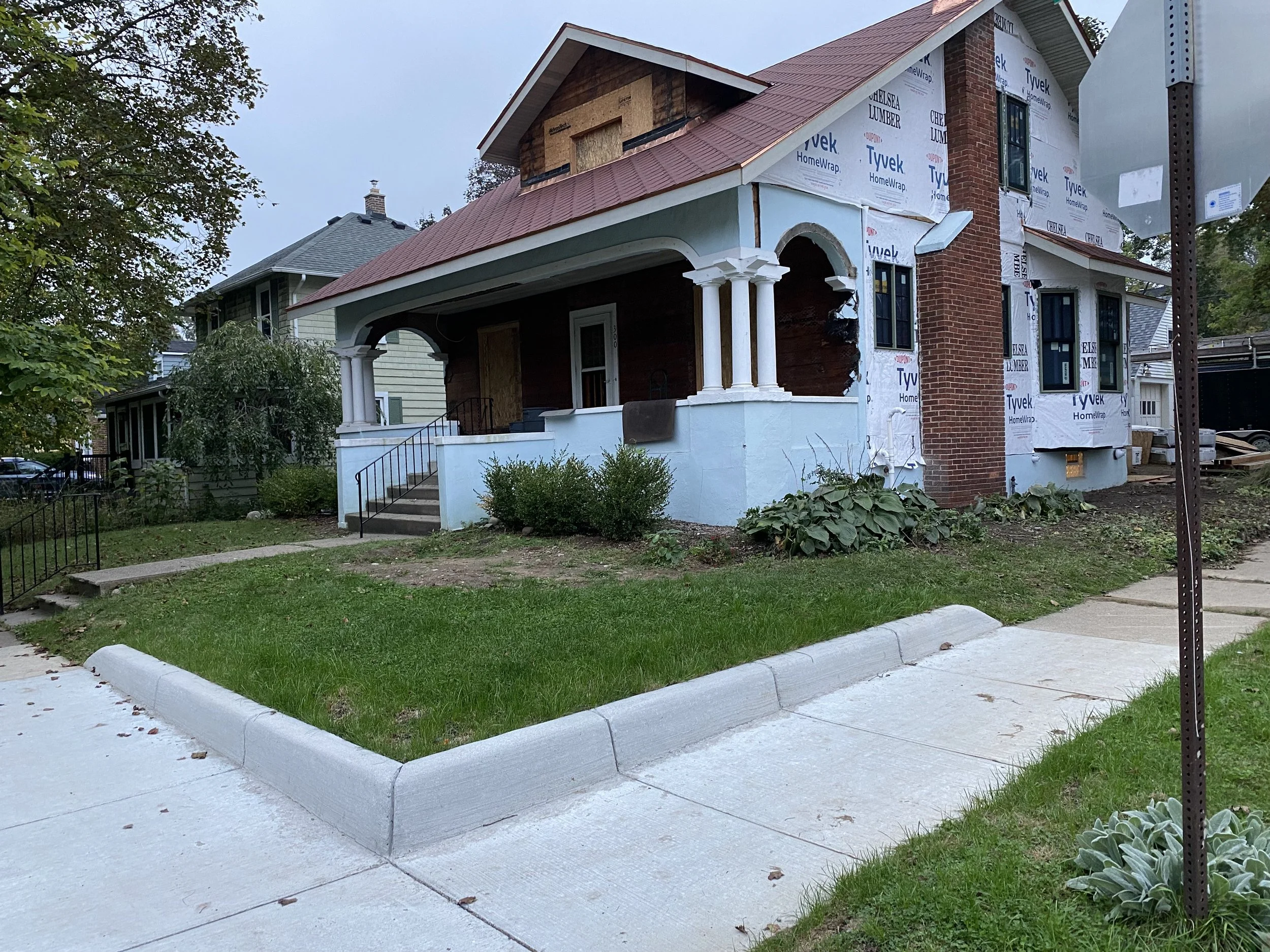 Front corner before the project — the house mid-renovation with a plain concrete curb and retaining wall meeting the sidewalk