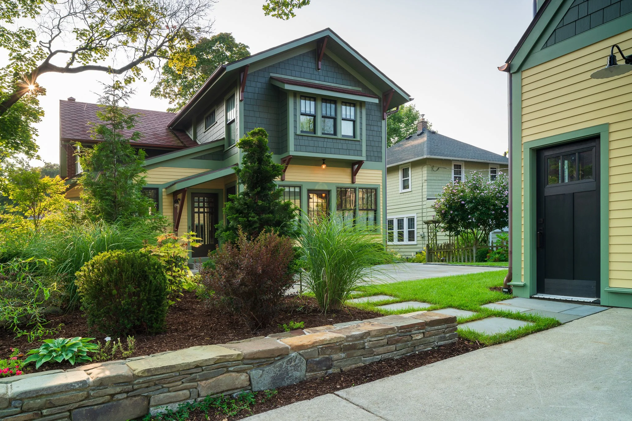 Front yard garden with shrubs, flowers, and a stone border in front of a sidewalk, with a two-story house with green siding and large windows in the background.