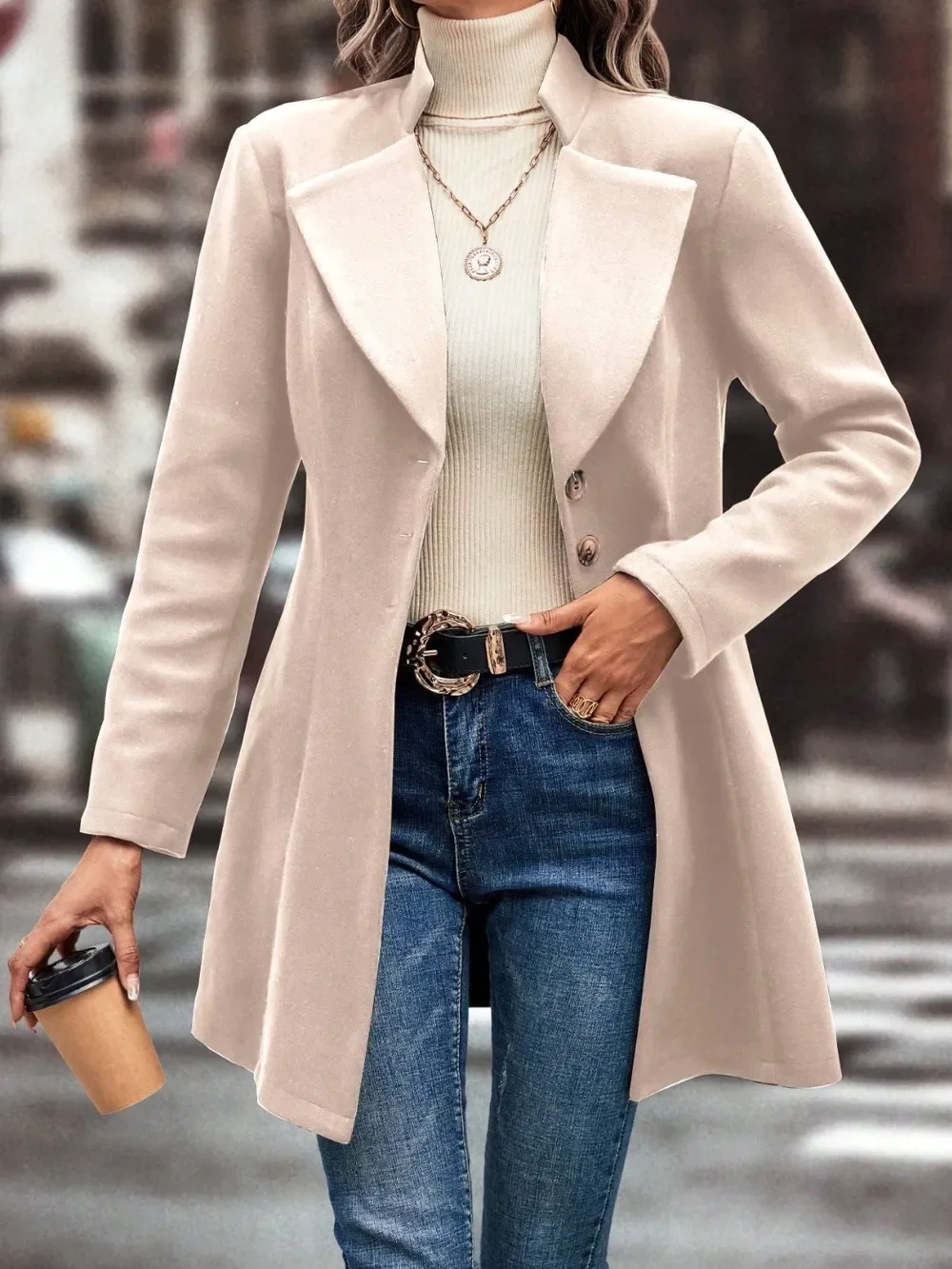 Woman wearing a beige coat over a light turtleneck sweater, blue jeans with a belt, holding a coffee cup in her left hand, on a city street.