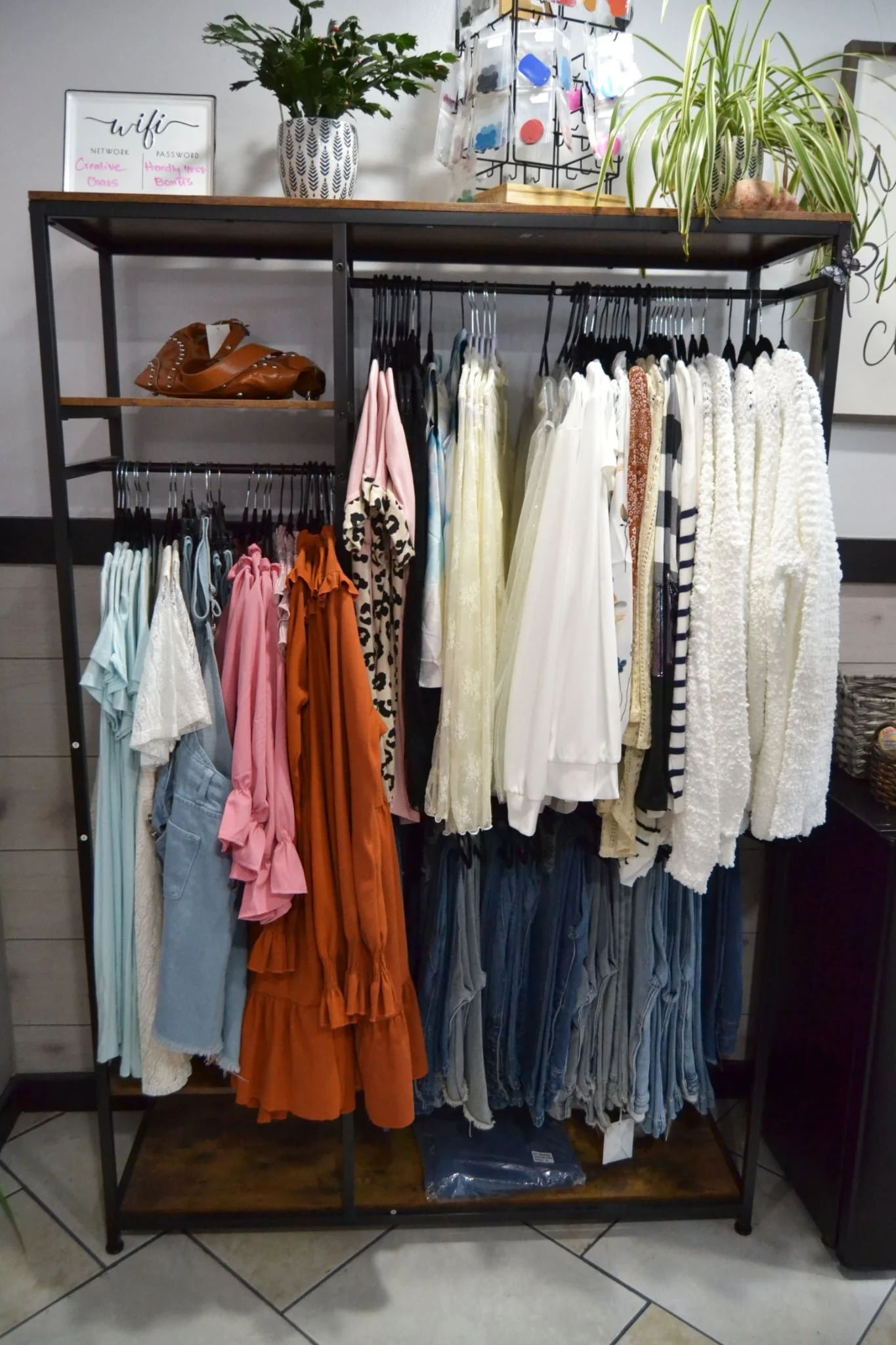 Clothing rack with various women's garments in pastel and neutral colors, including dresses, tops, and sweaters. There are some plants and decorative items on top of the rack, and a small whiteboard with Wi-Fi information visible in the background.