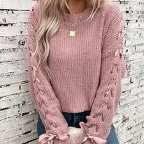 Woman wearing a pink knitted sweater with cut-out details on the sleeves, standing in front of a white brick wall.