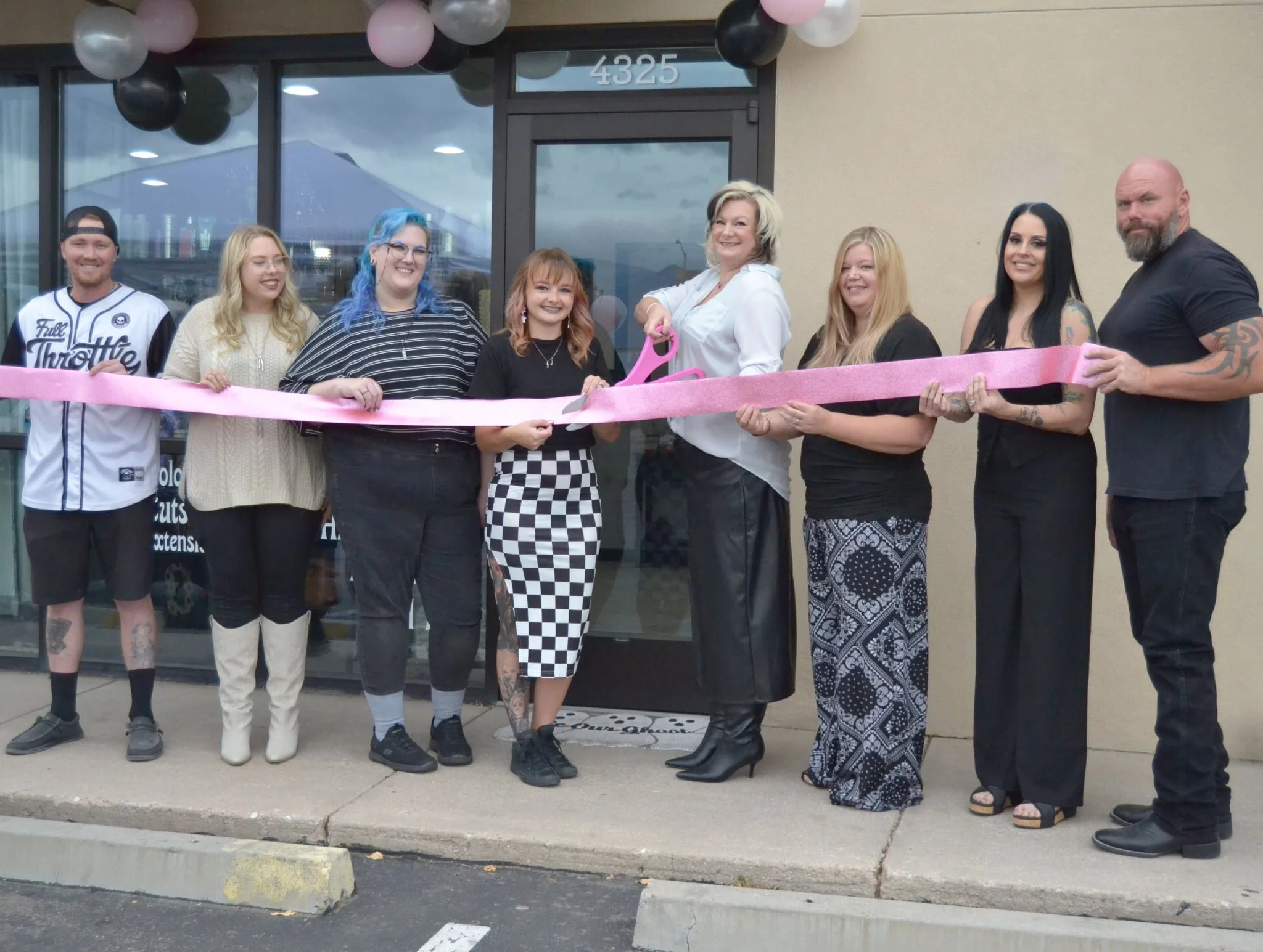 Group of people standing outside a building with balloons, holding a pink ribbon during a ribbon-cutting ceremony.