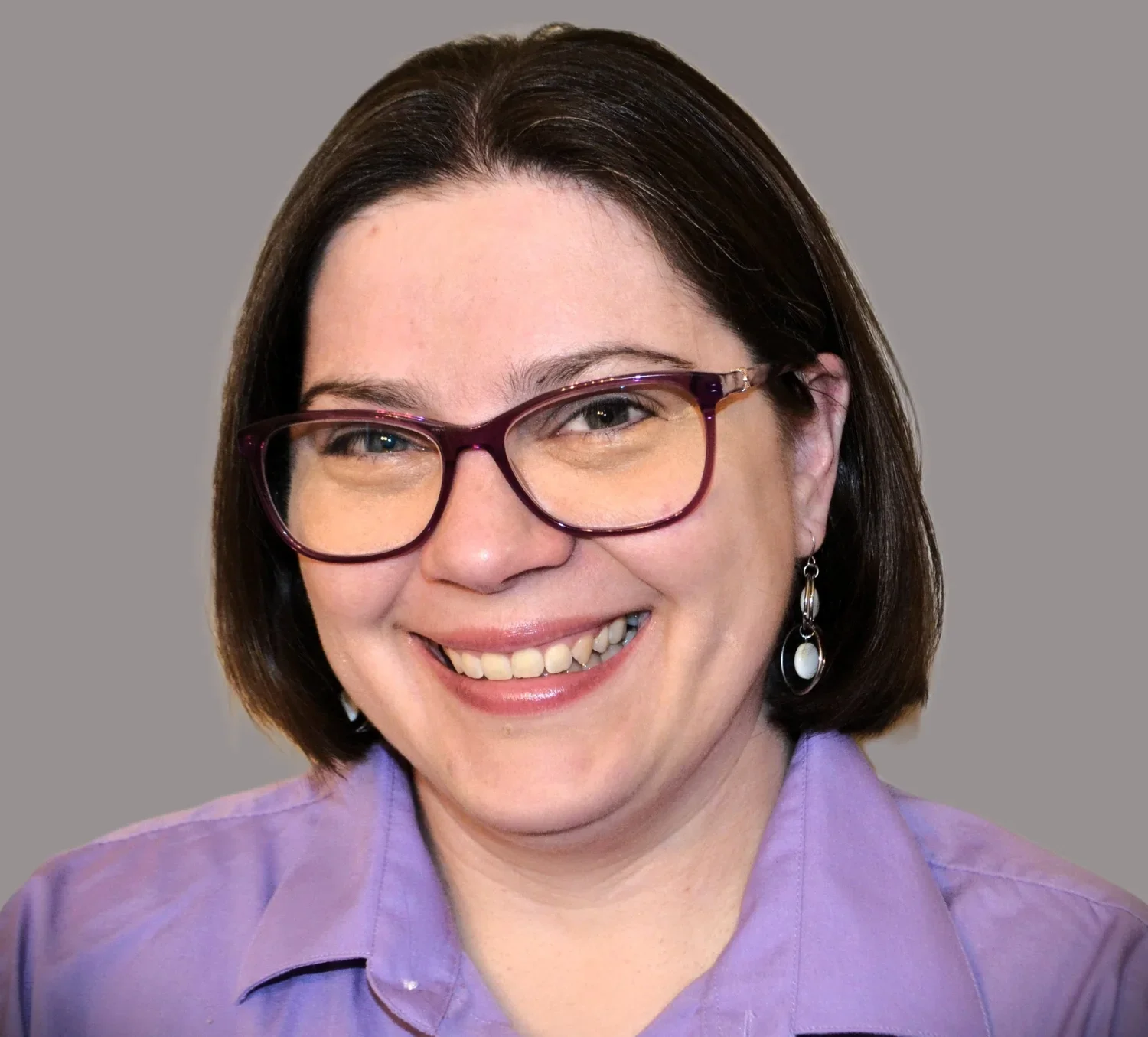 Portrait of a woman with shoulder-length dark hair, wearing glasses with a purple frame, silver earrings, and a lavender collared shirt, smiling against a plain gray background.