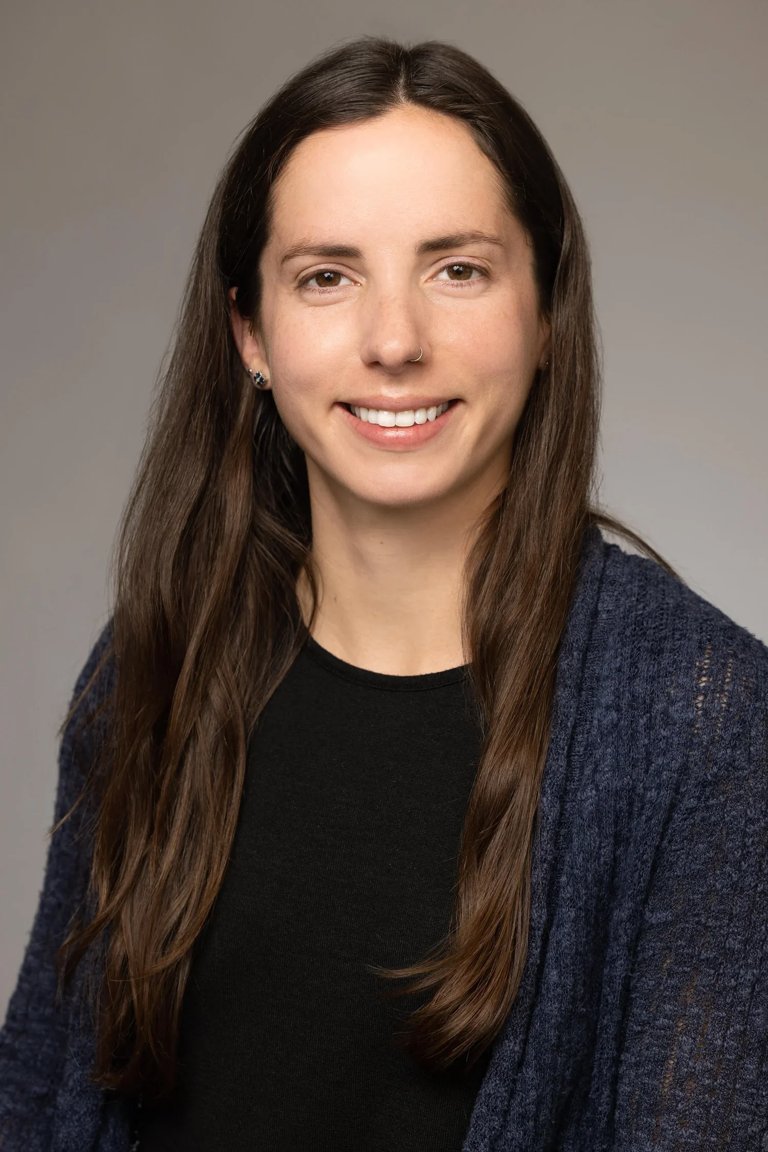 Headshot of a woman with long brown hair, smiling, wearing a black top and a dark blue cardigan, against a plain gray background.