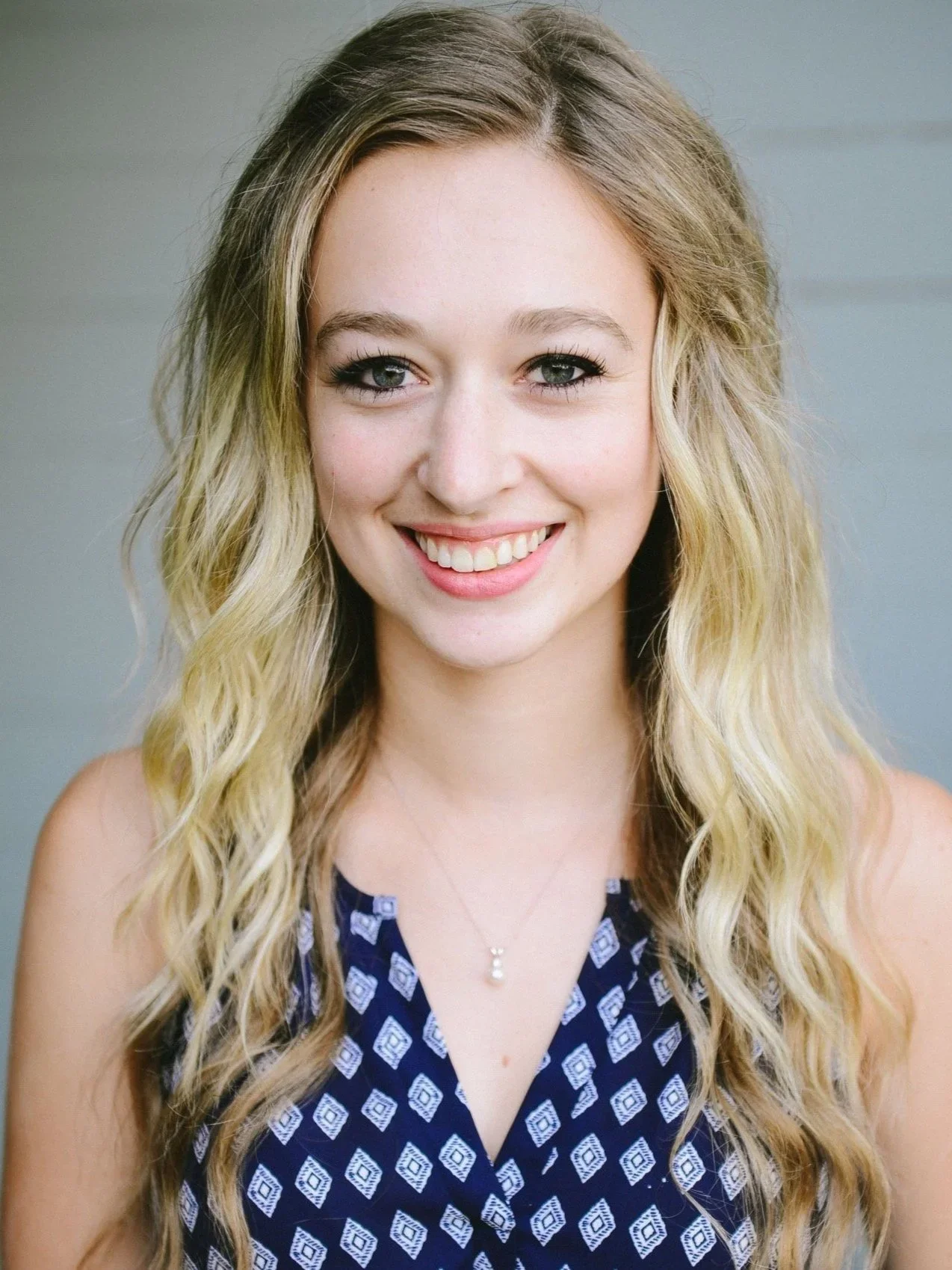 Close-up portrait of a young woman with long, wavy blonde hair, blue eyes, and a bright smile, wearing a navy blue patterned sleeveless top and a delicate necklace, against a neutral background.
