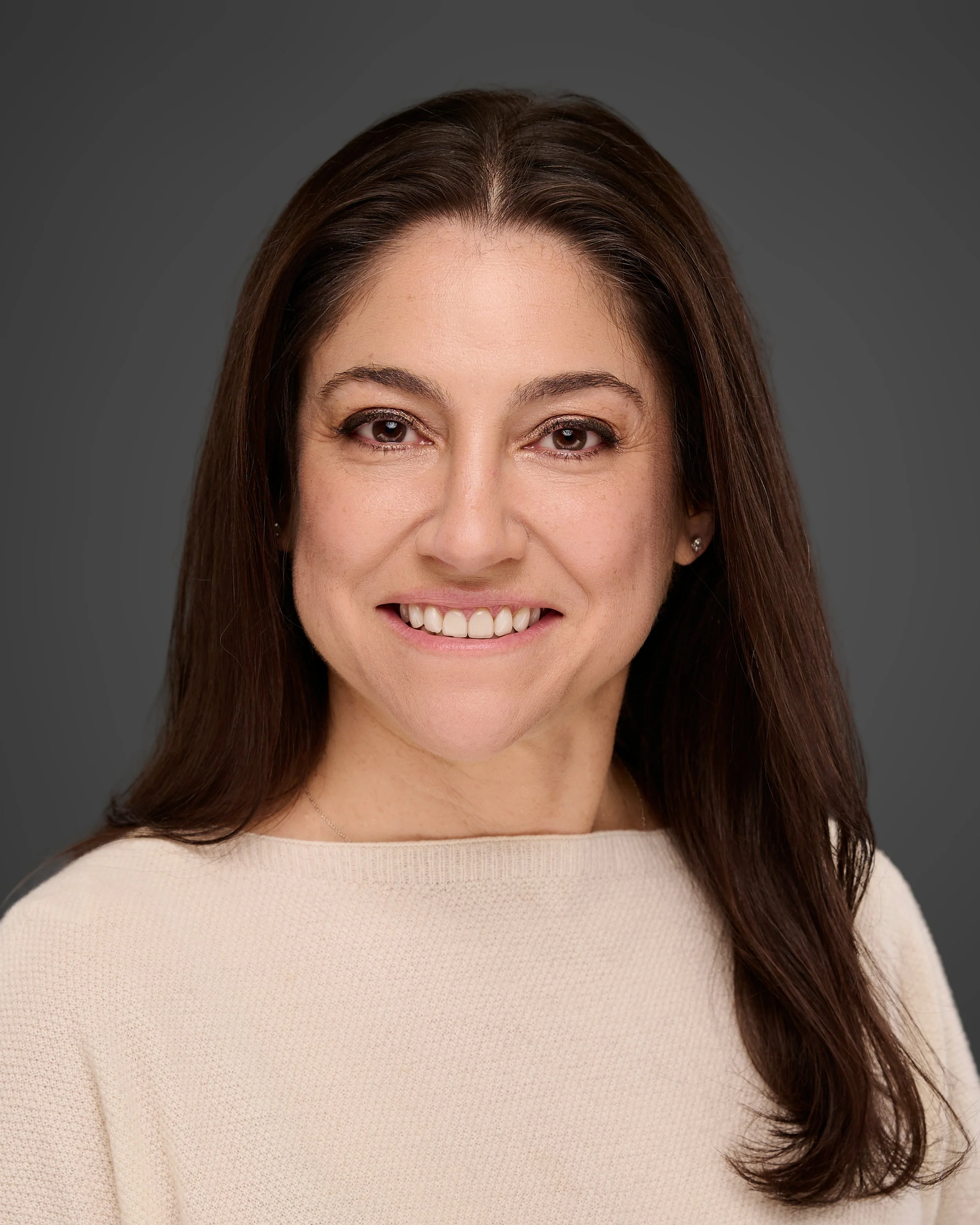 A portrait of a woman with dark brown hair and brown eyes, wearing a sleeveless purple shirt and a subtle smile, against a neutral gray background.