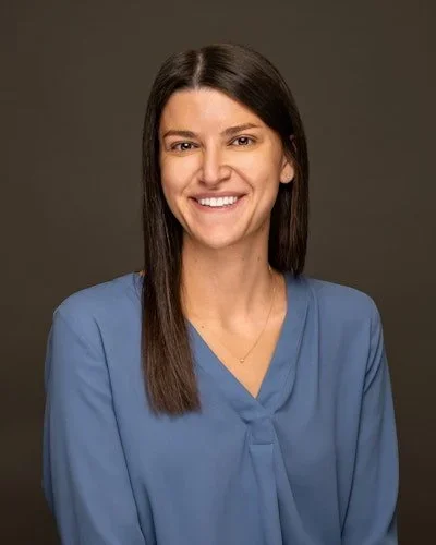 A woman with long dark hair smiling, wearing a blue blouse, against a plain dark background.