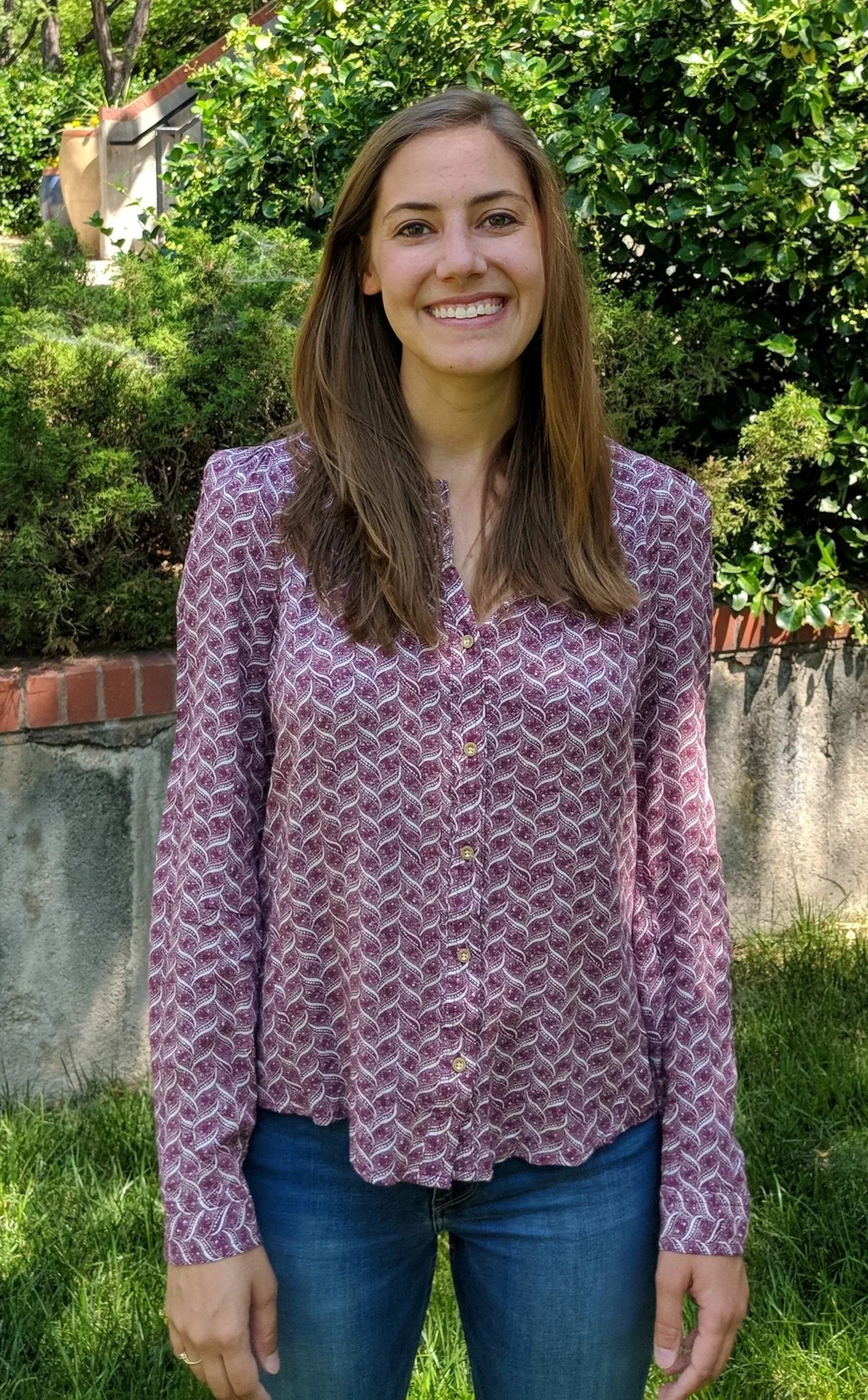 A young woman with long brown hair smiling outdoors, wearing a patterned purple blouse and blue jeans, standing in front of green bushes and trees.