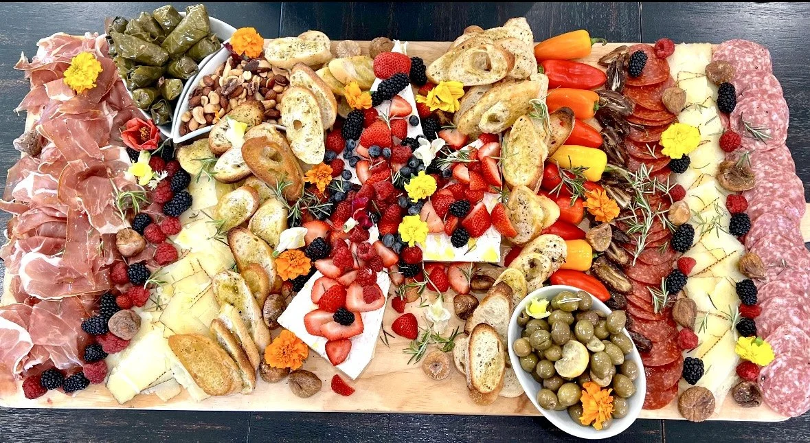 A large charcuterie board with assorted cured meats, cheeses, crackers, grapes, strawberries, blackberries, raspberries, cherry tomatoes, pickles, and garnished with edible flowers and herbs.