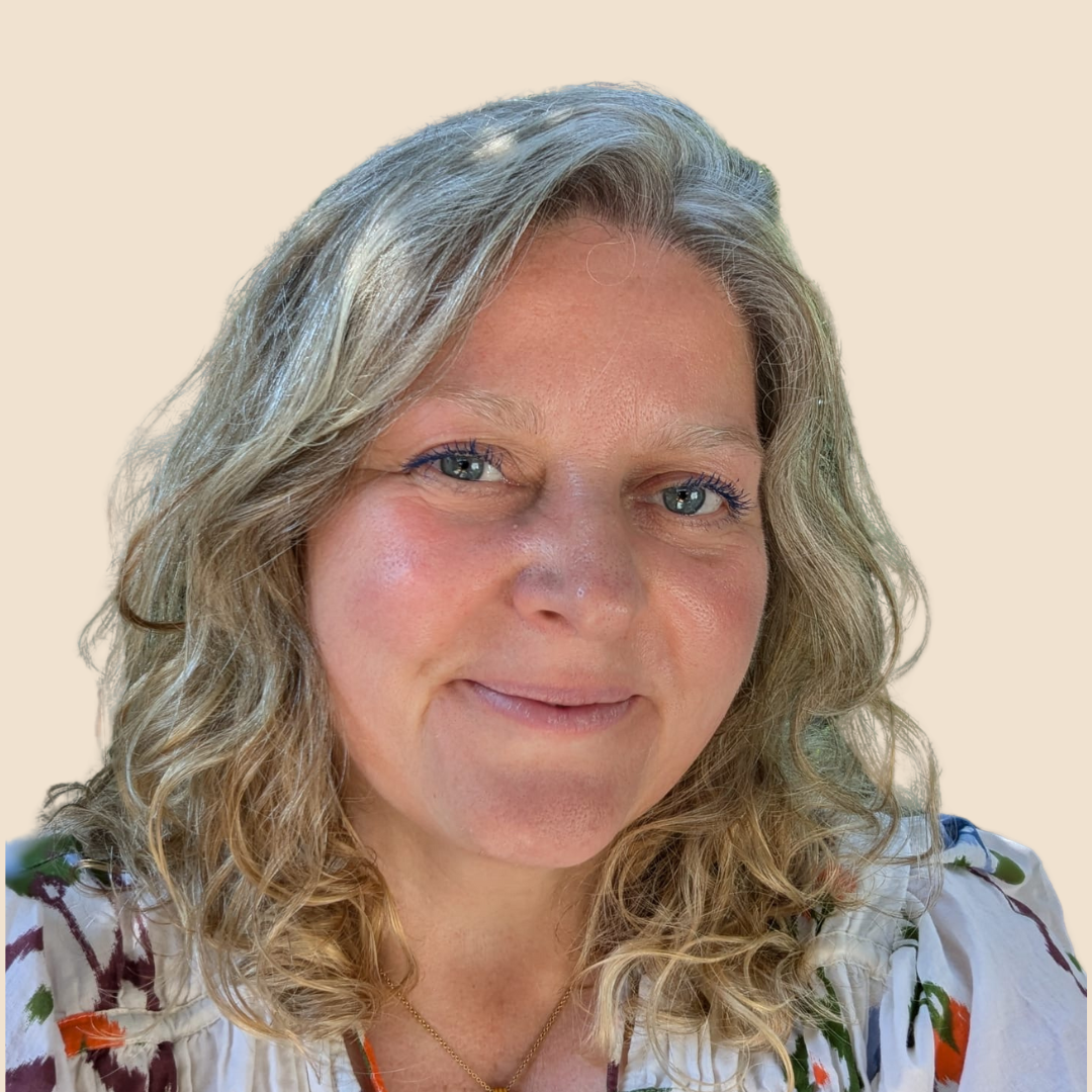 Close-up of a woman with wavy blonde hair, blue eyes, light skin, and a subtle smile, wearing a white top with colorful floral patterns and a small gold necklace, against a plain light background.