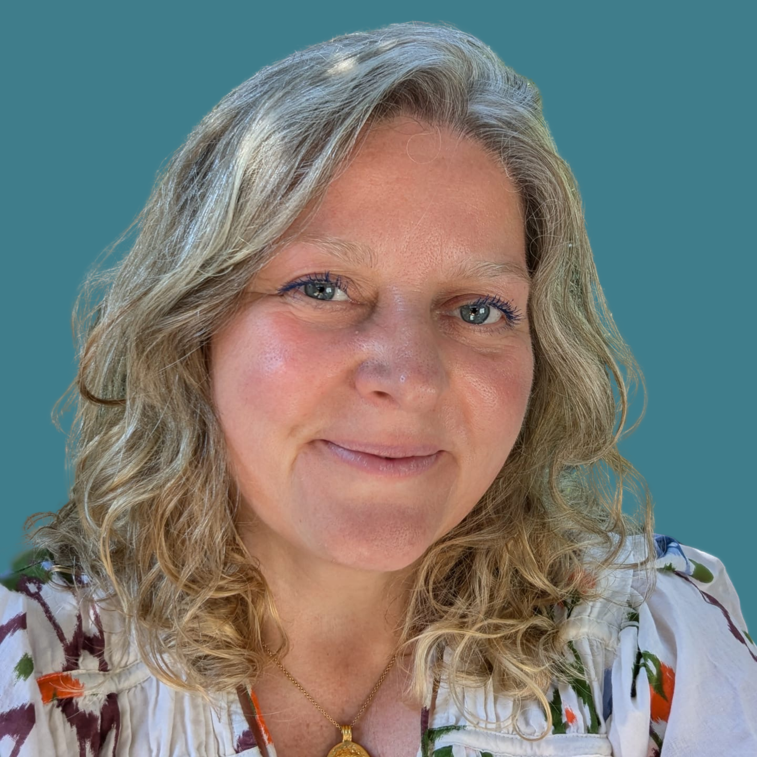 Clare Cusack smiling gently, wearing a white top with a subtle floral pattern, against a teal background.