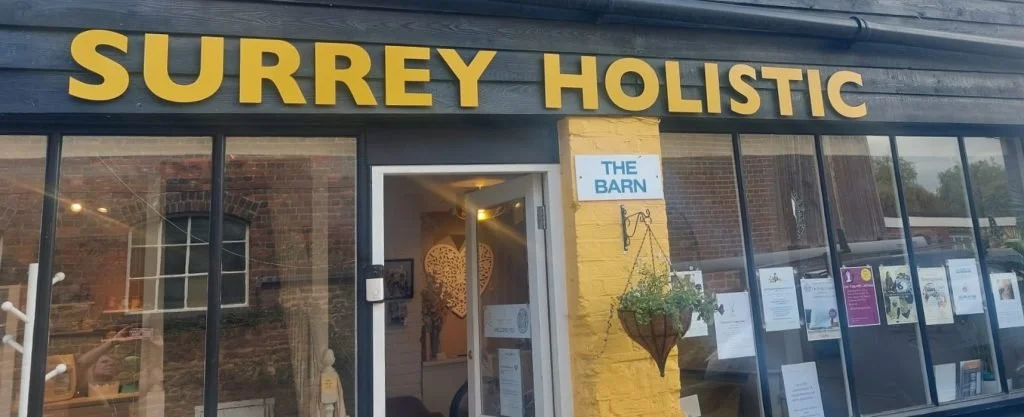 Storefront of Surrey Holistic, a holistic health center with glass windows displaying informational posters, a hanging plant outside, and a sign indicating 'The Barn' entrance.