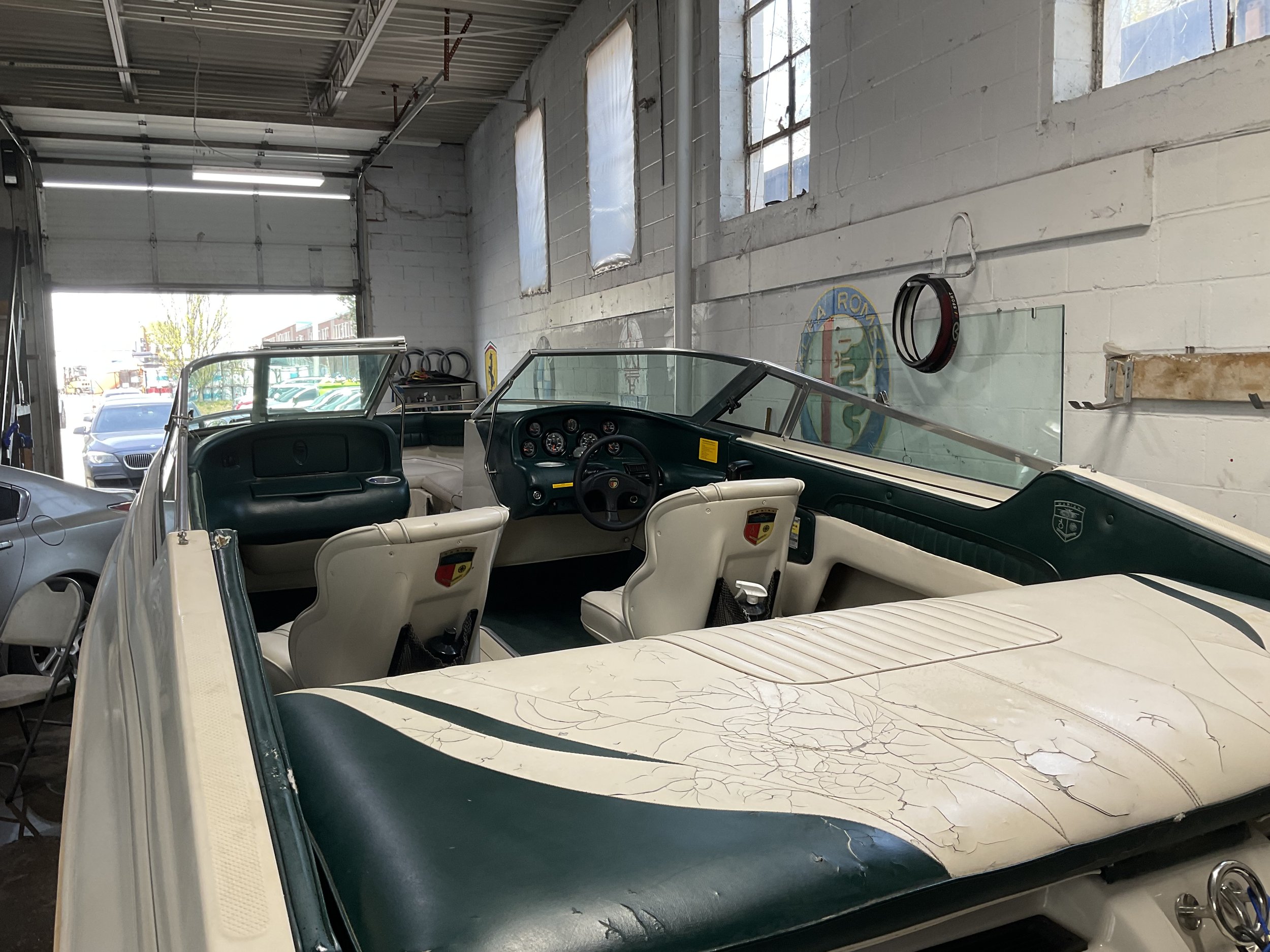 Interior view of a vintage boat with white and dark green seats, steering wheel, and dashboard inside a garage.