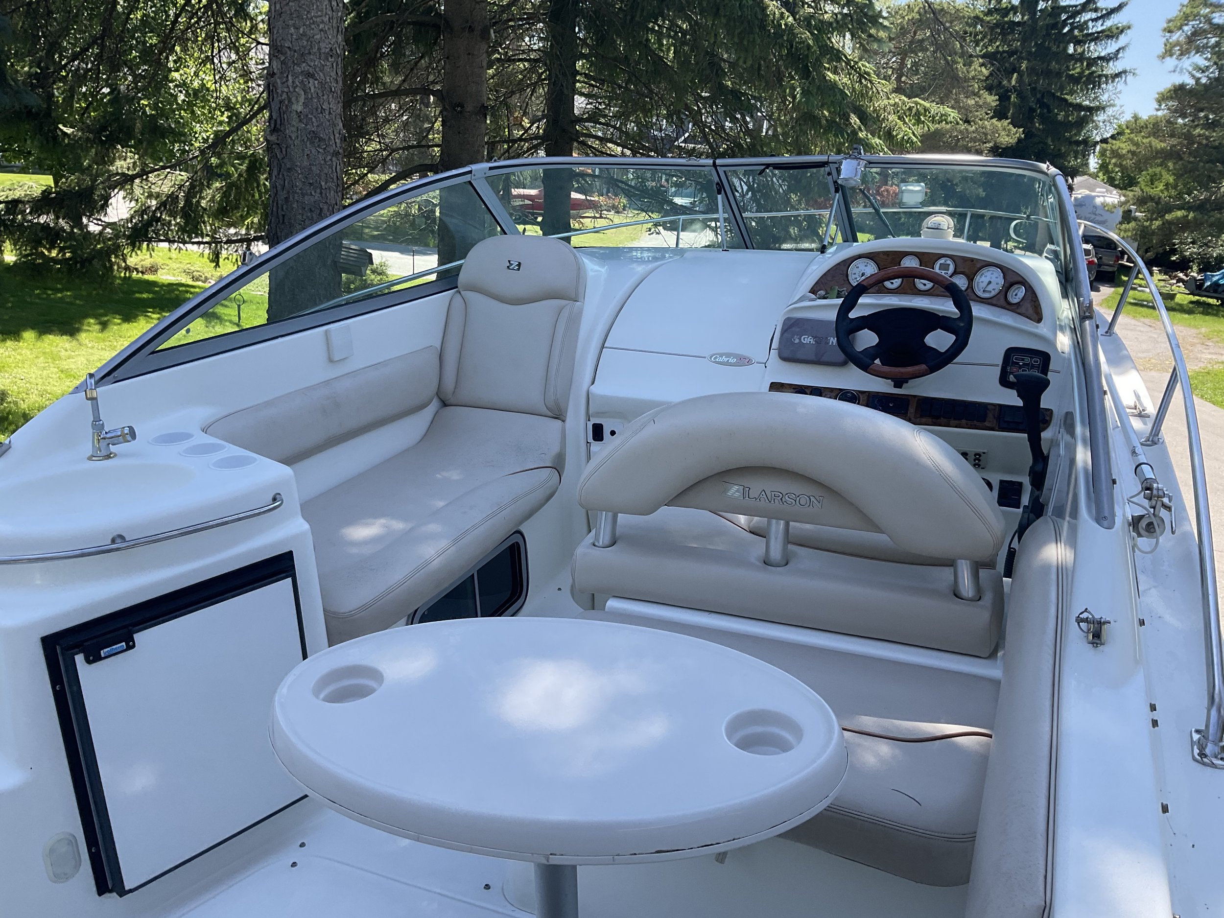 Interior of a Larson boat, featuring a steering wheel, dashboard, white cushioned seating, a small round table with cup holders, and a compact kitchenette with a sink.