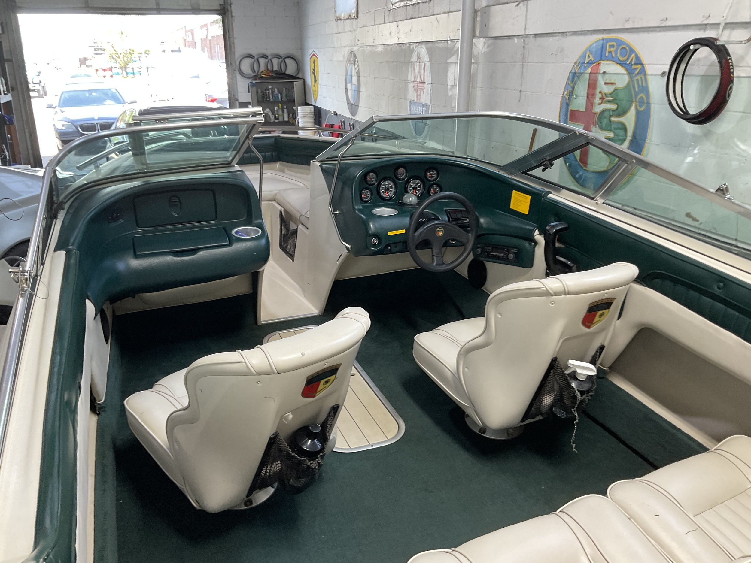 Interior of a motorboat parked in a garage, featuring two white leather seats, green carpet, a dashboard with gauges and a steering wheel, and car logos on the wall.