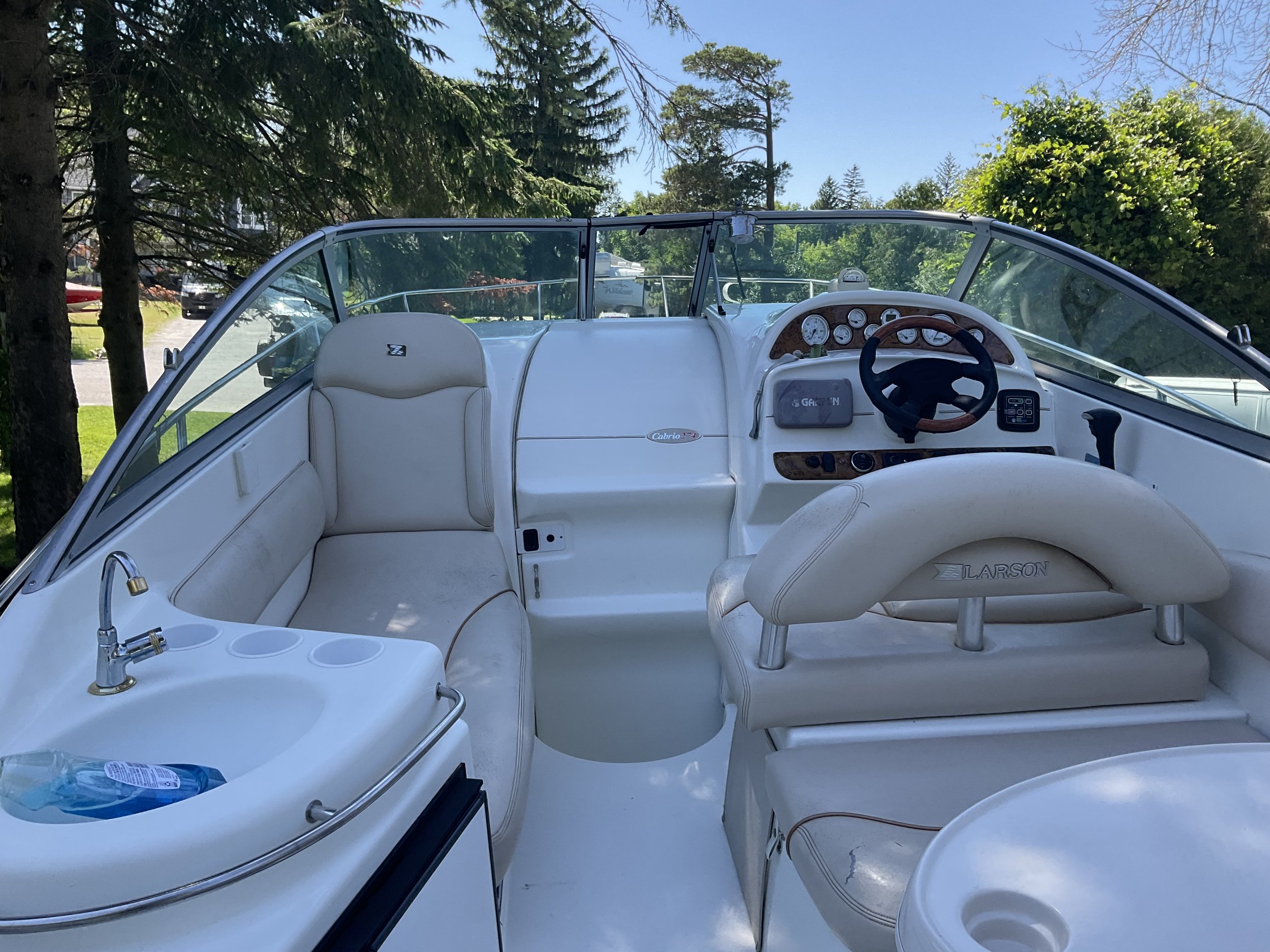 Interior of a white Larson motorboat with seats, a steering wheel, control panel, sink, and cup holders, surrounded by trees.