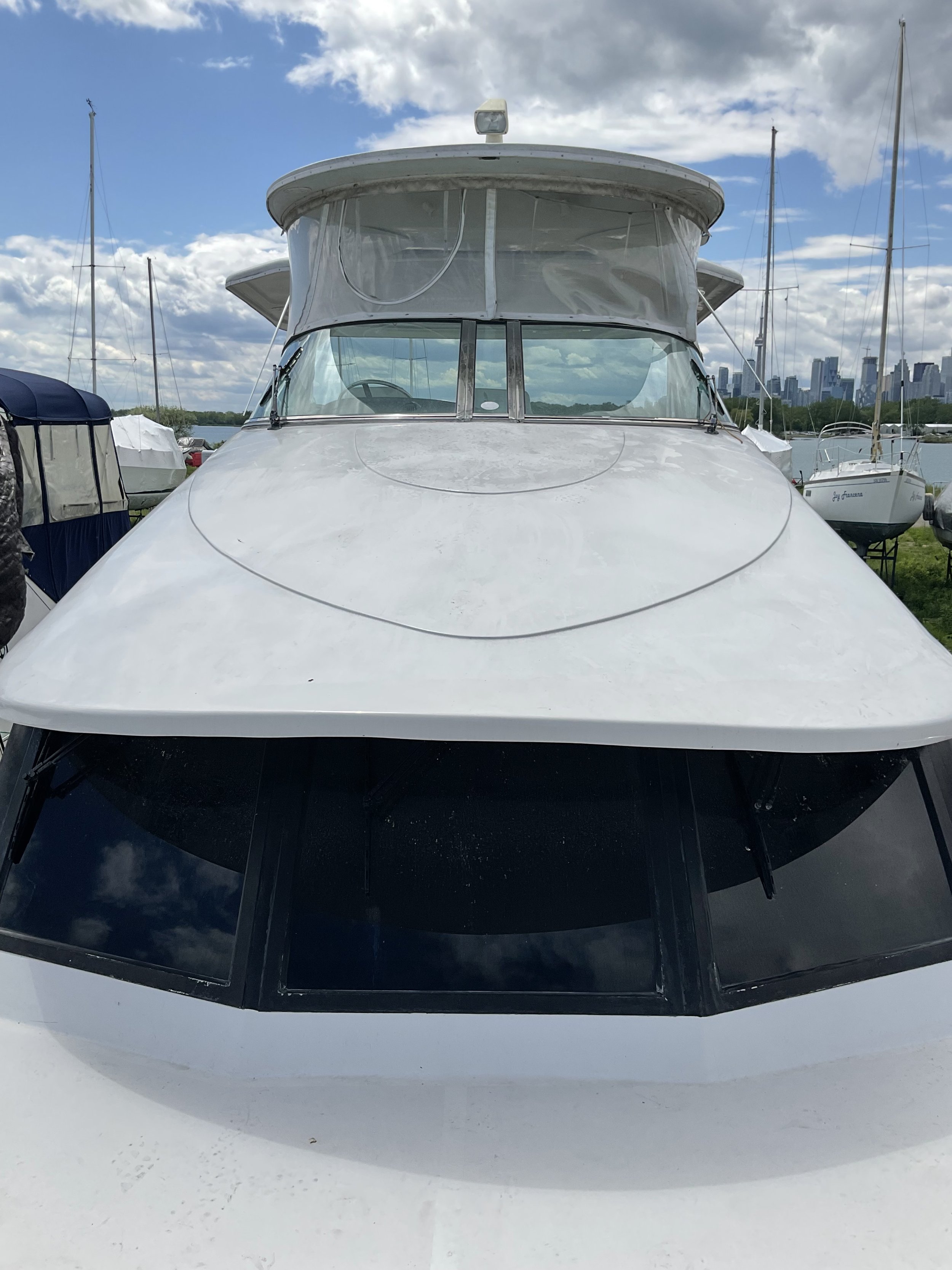 Close-up of a white yacht with a visible windshield and canopy, set against a marina background with sailboats and a city skyline under a cloudy sky.
