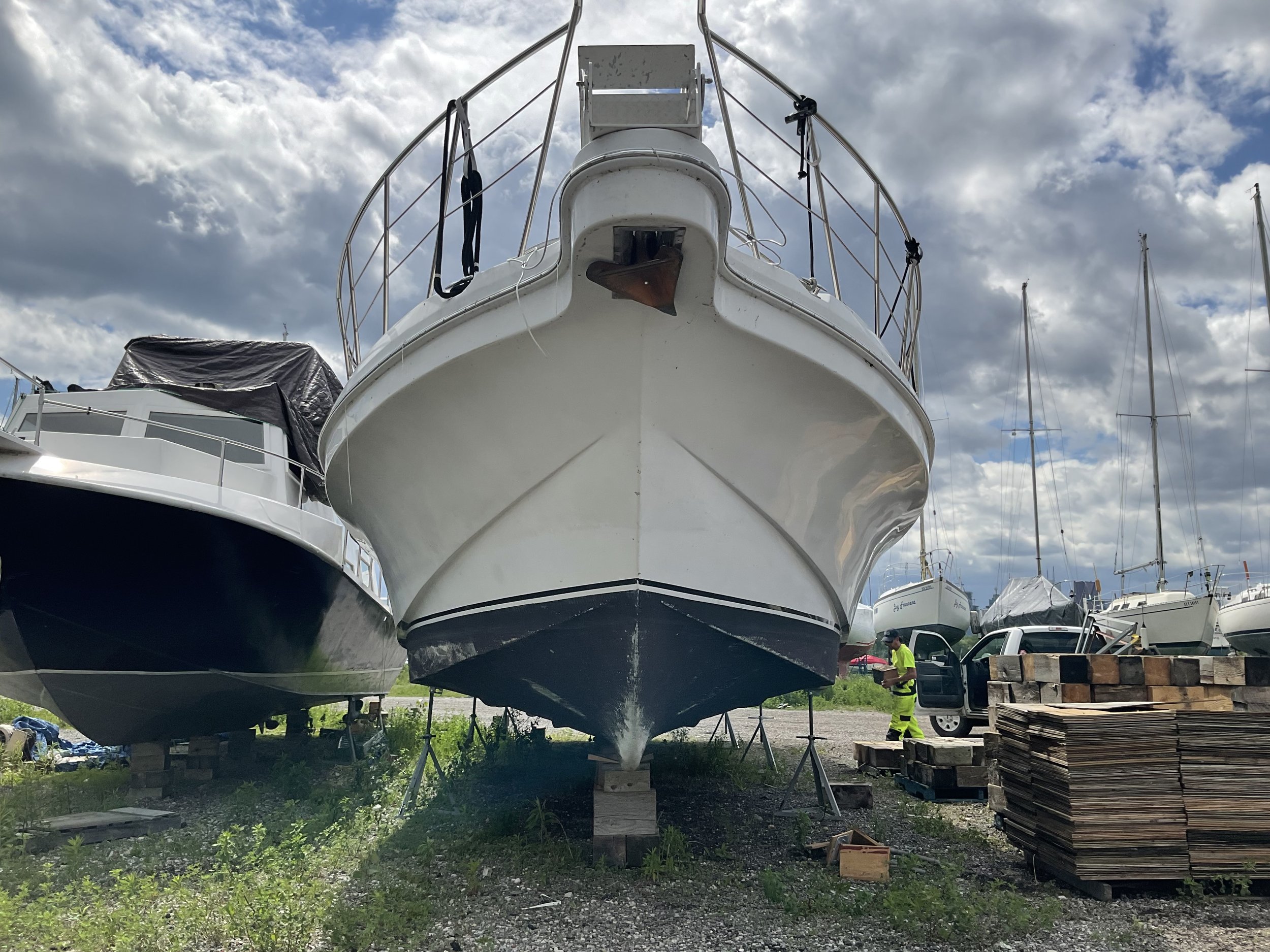 Front view of two boats on land, with a cloudy sky and several other boats in the background. A person is visible near a truck on the right, with piles of wooden planks nearby.