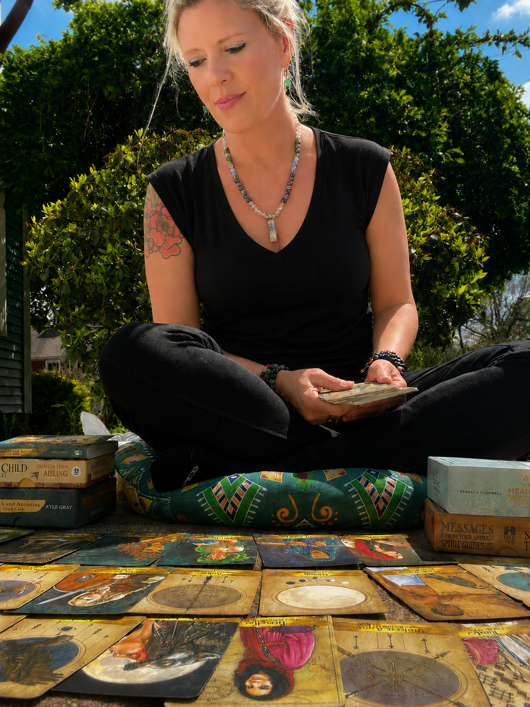 A woman sitting outdoors on a cushion, looking at tarot and oracle cards spread on the ground in front of her, with a lush green bush and blue sky in the background.