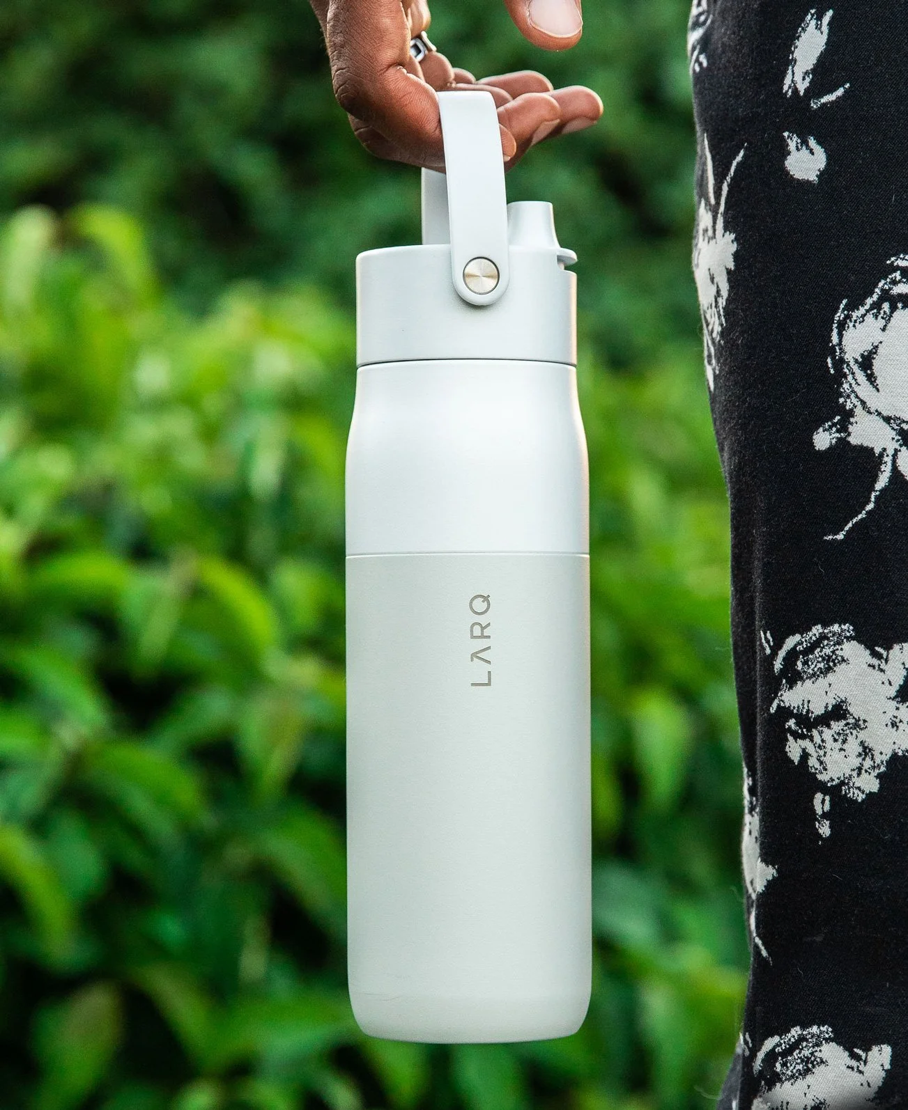 A person holding a white LARQ portable water bottle outdoors with green foliage in the background.