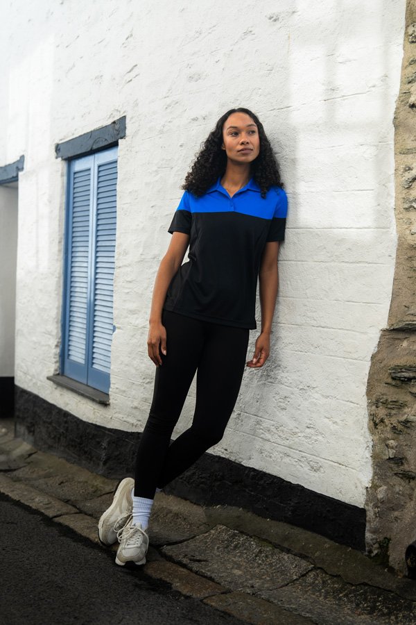 A woman with curly black hair standing against a white brick wall near a blue window shade, wearing a black and blue sports shirt, black leggings, white sneakers, and white sock, on a cobblestone street.