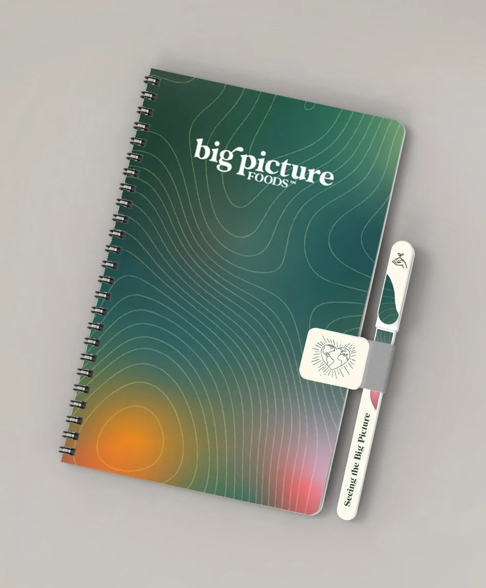 A spiral notebook with a green cover titled 'big picture FOODS' and a matching band, accompanied by a pen that has 'Seeing the Big Picture' engraved on it, placed on a light grey surface.