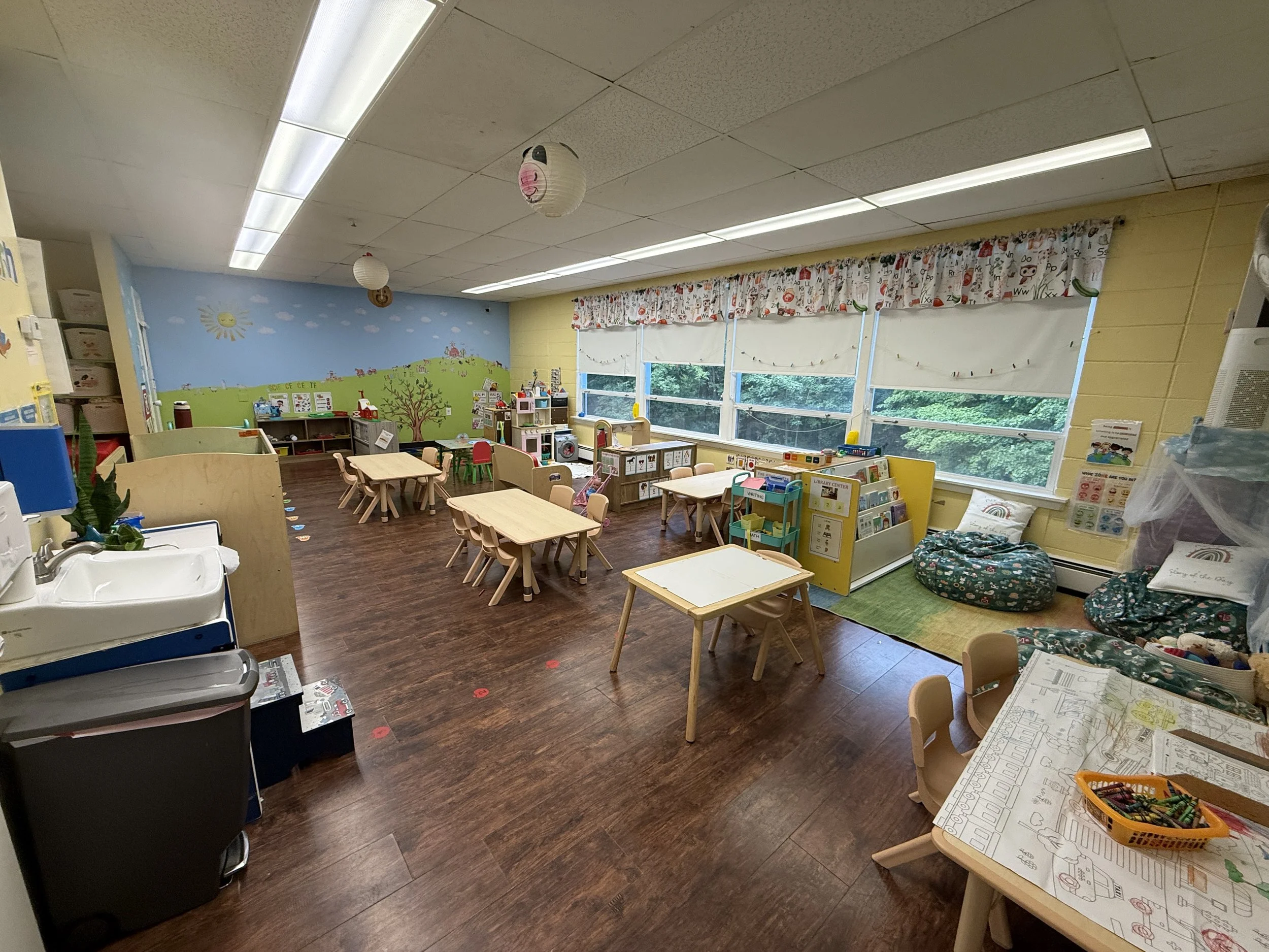 A classroom with small tables and chairs, a colorful mural on the wall, and windows with curtains. There are various educational materials and toys around at Garden Grove Preschool in Sparta NJ