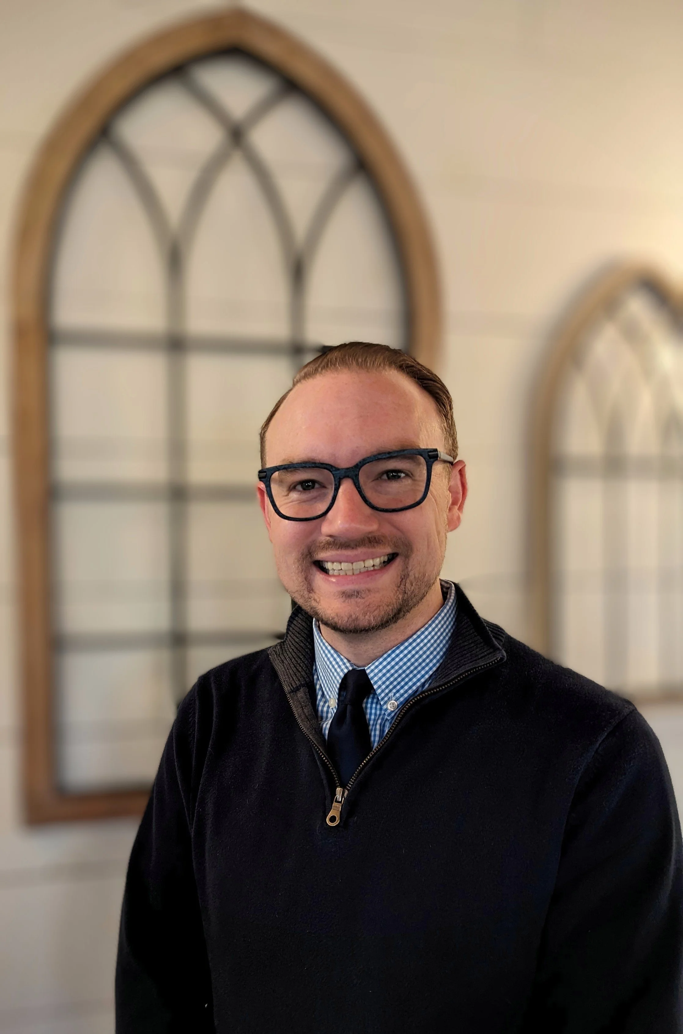 A man wearing glasses, a blue checkered shirt, a black tie, and a black zip-up sweater, smiling in front of a blurred background of two arched windows.
