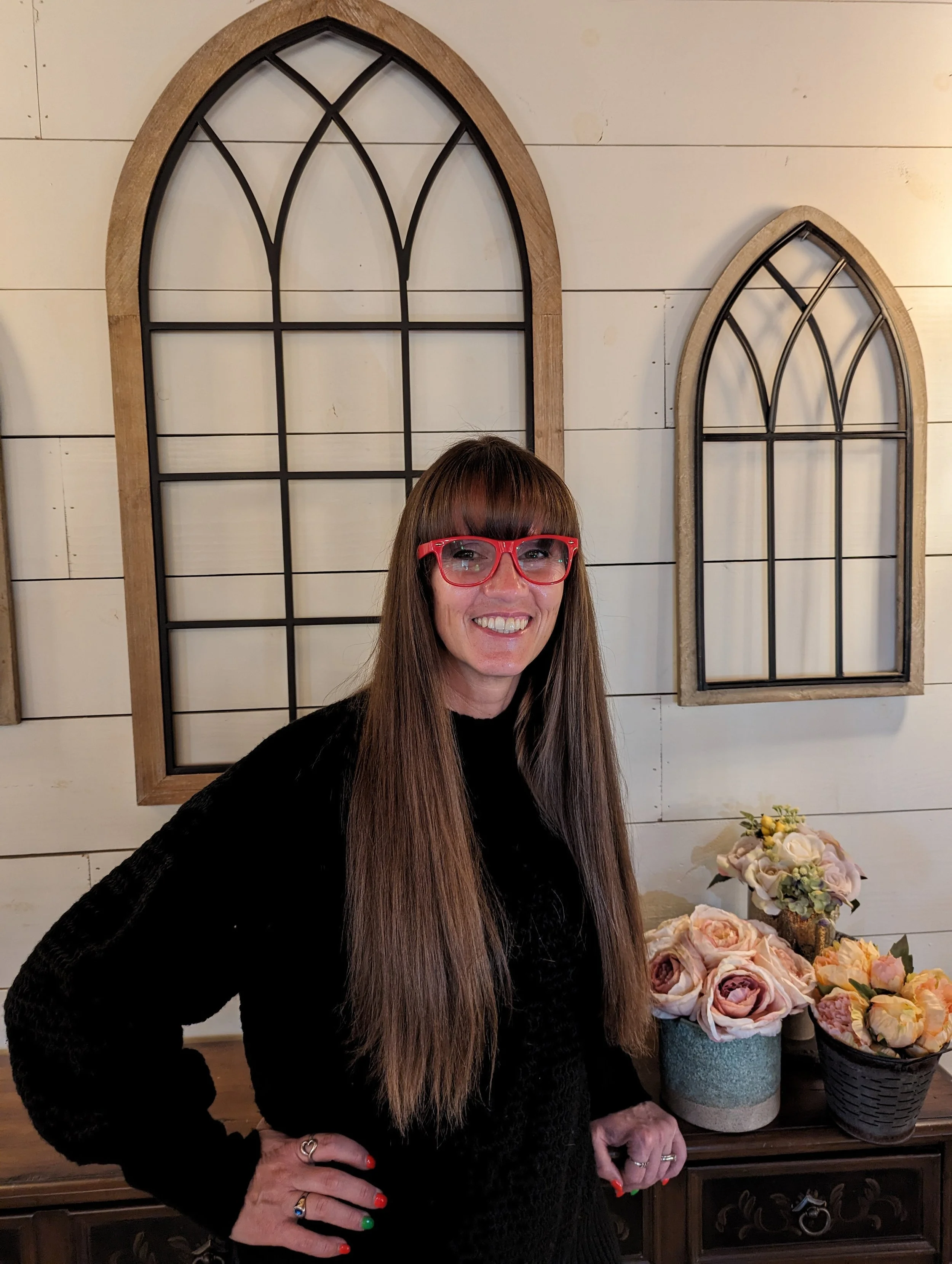 A woman with long brown hair and red glasses smiling while standing in front of a wall with decorative window frames and a table with flower arrangements.