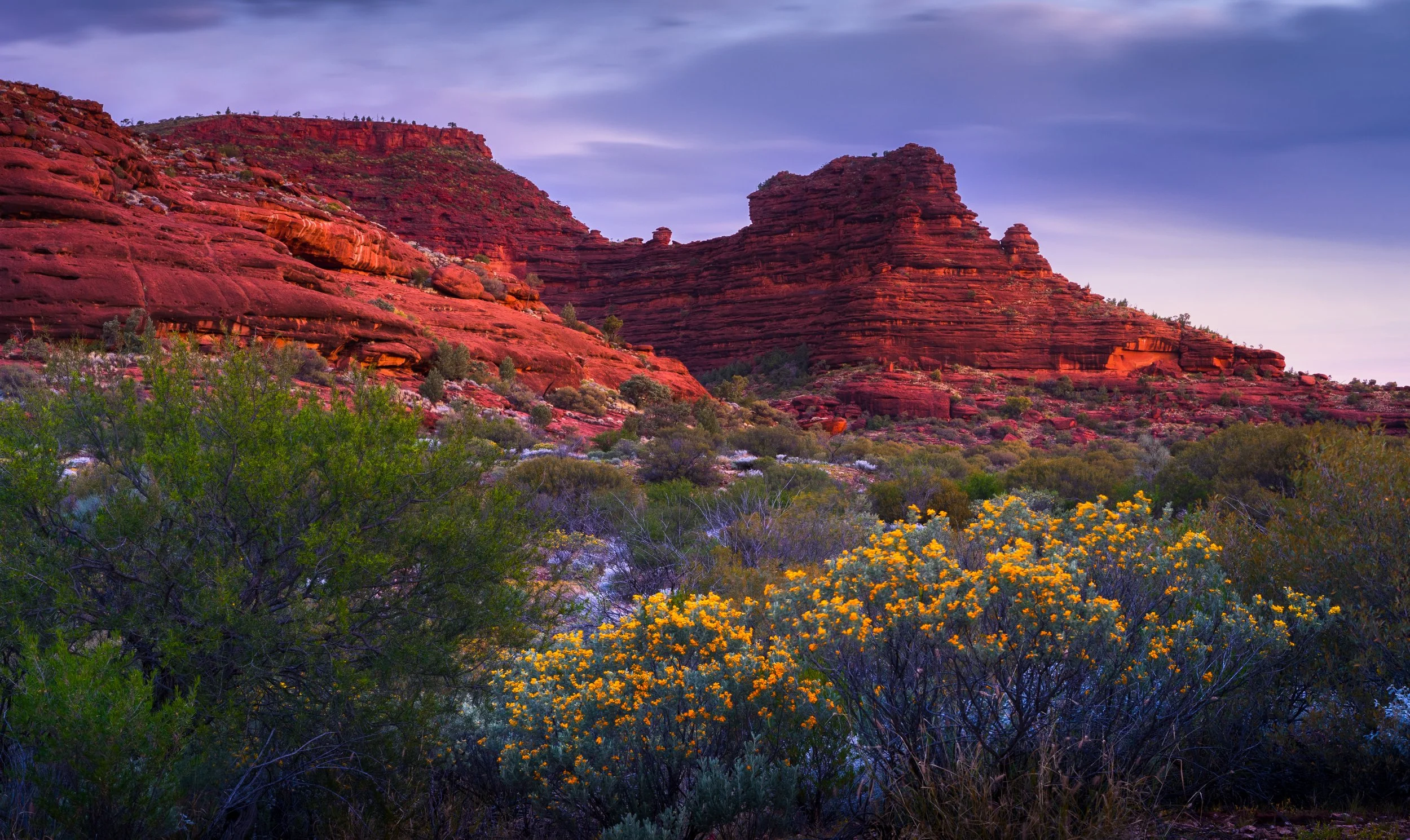 Red rock formations with layered cliffs and a shrub-filled foreground, under a partly cloudy sky at sunset.