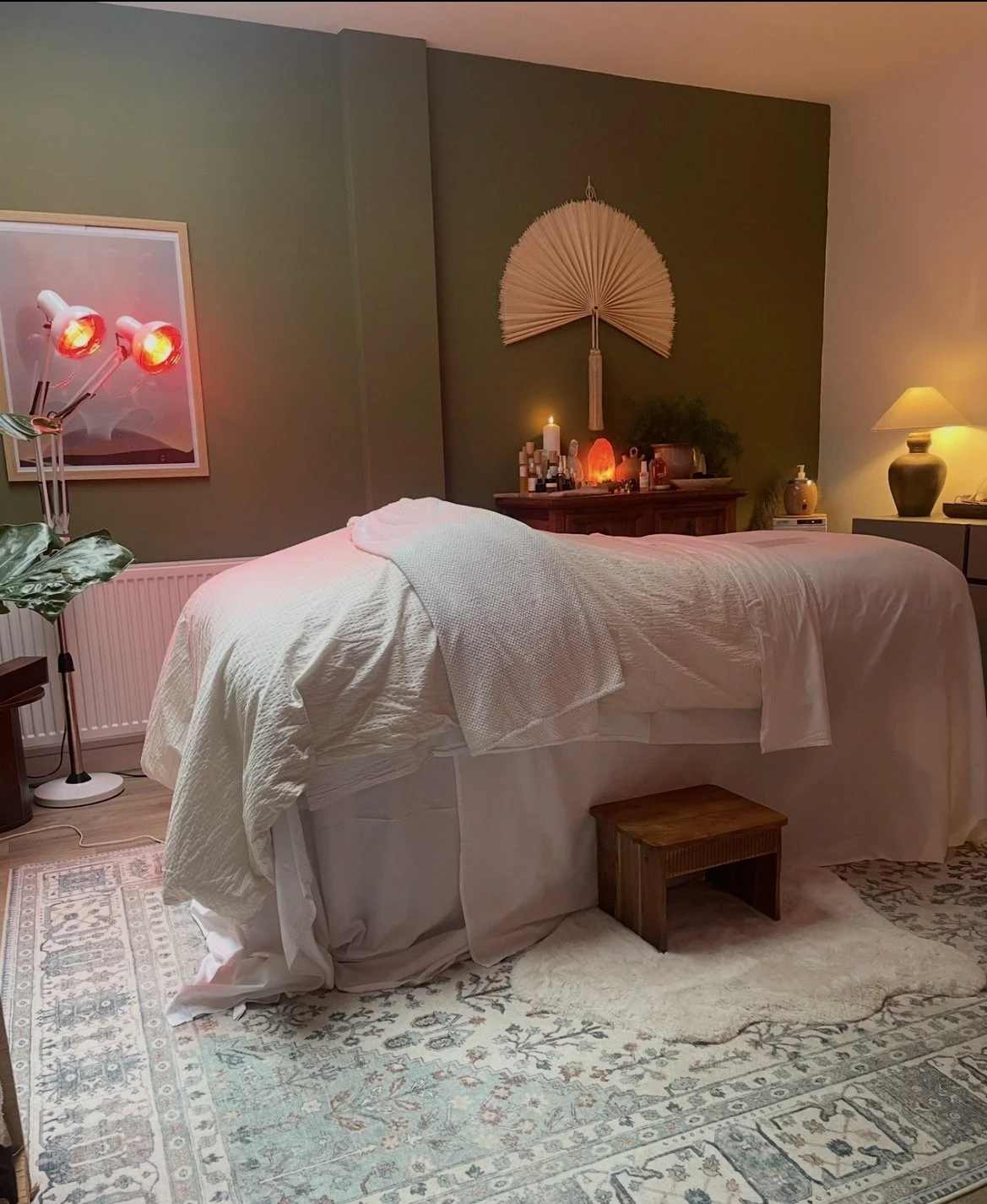 A massage or therapy room with a bed covered in white sheets, dimly lit with warm and red lighting, decorated with candles, plants, and spiritual items.