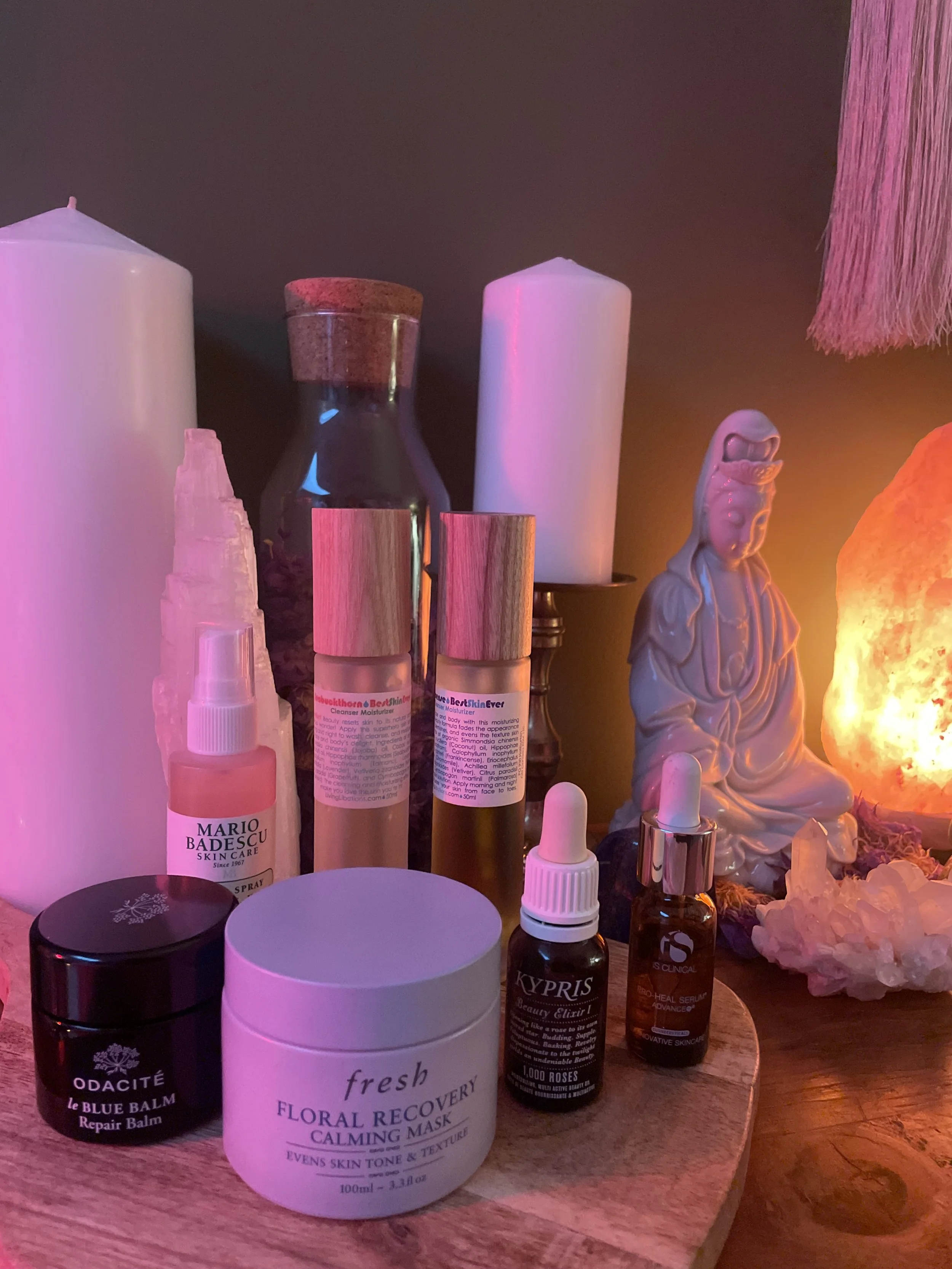 A collection of skincare products, candles, and decorative objects on a wooden surface, illuminated by warm pink and orange lighting.