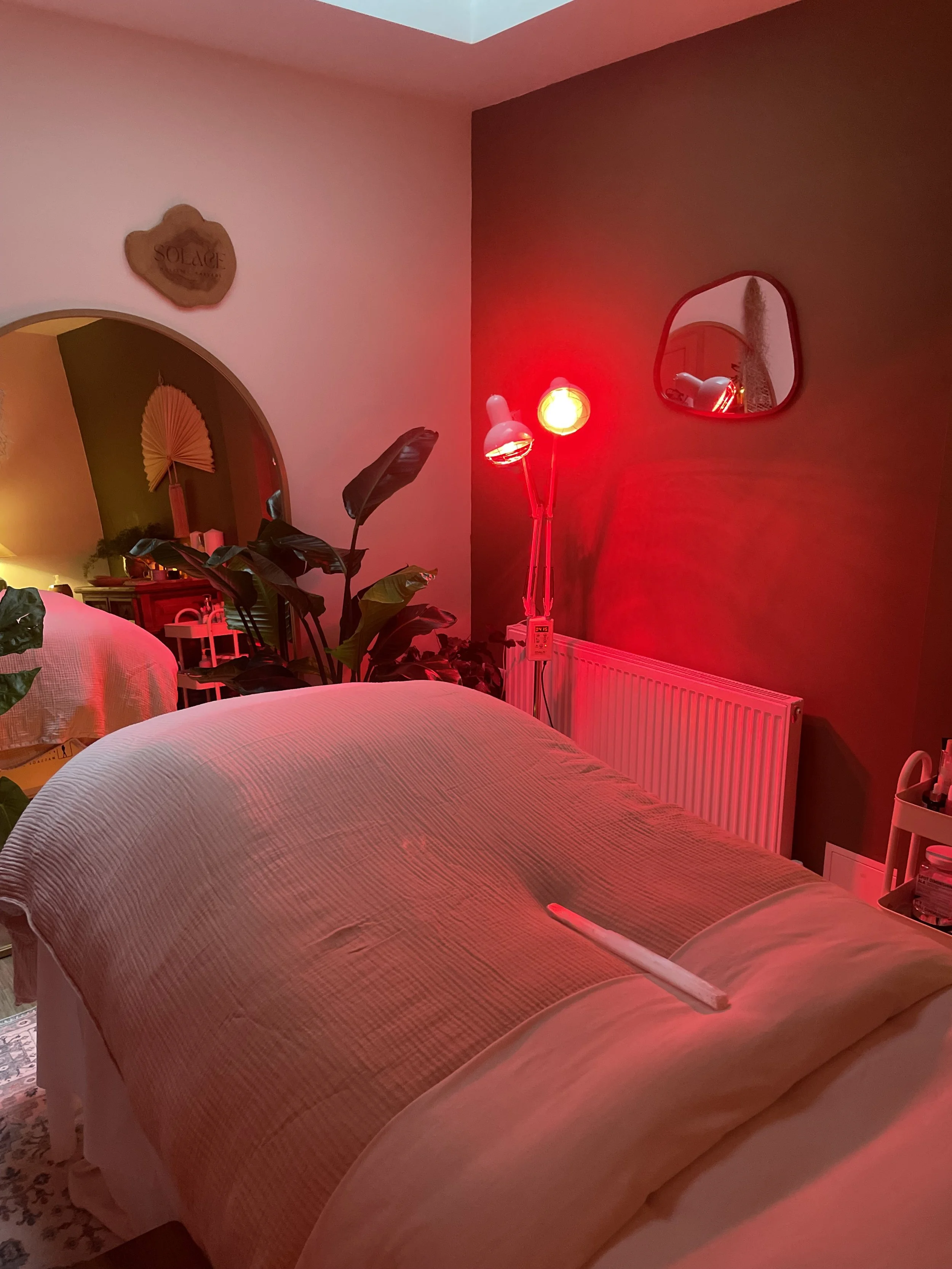 A cozy bedroom with warm lighting, a bed with a light-colored quilt, a standing lamp emitting red light, a mirror on the wall, plants near the window, and decorative items on shelves.