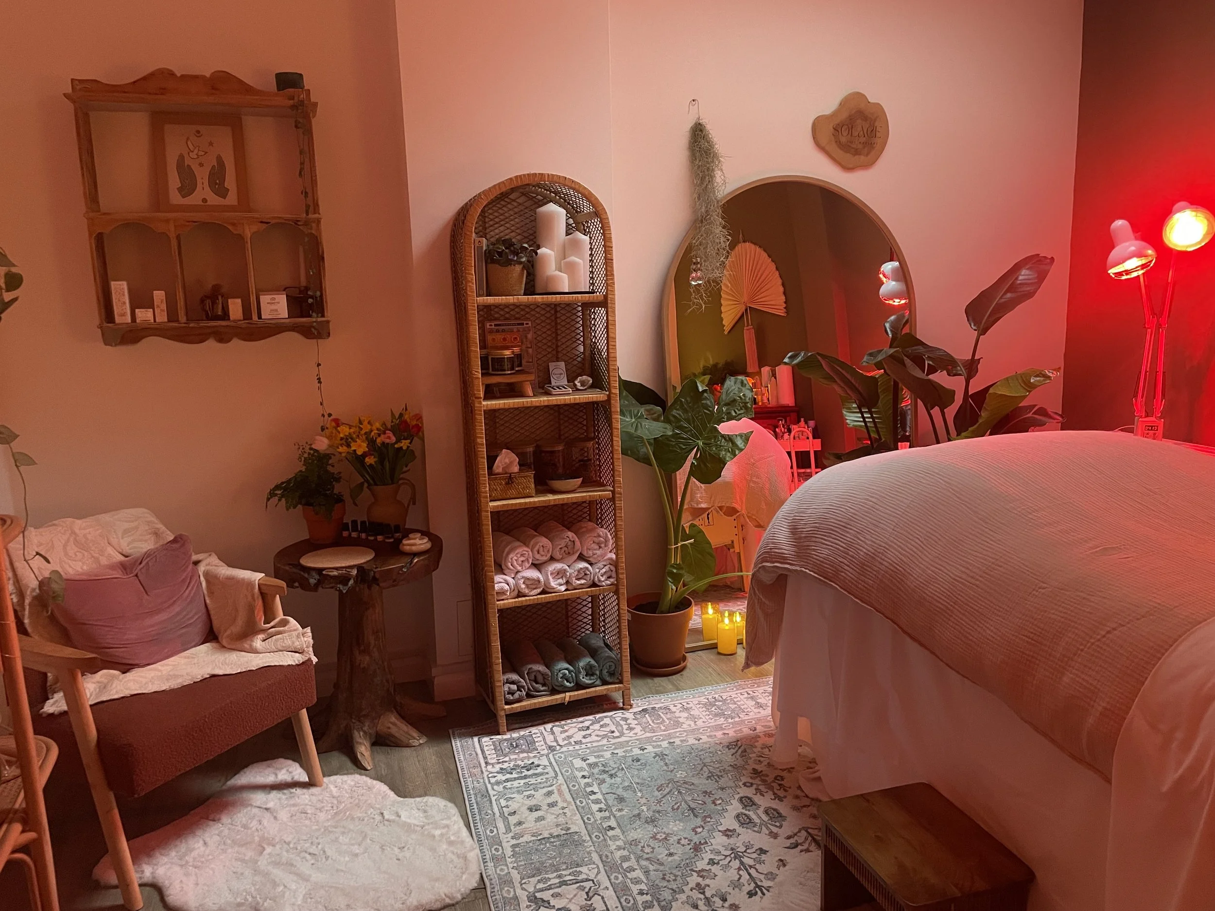A cozy bedroom decorated with warm lighting, plants, and shelving. Features a bed, a chair with cushions, a small round table with a flower pot, a wooden shelf with candles, towels, and decorative items, and a mirror reflecting the room. There are candles on the floor and an adjustable lamp with red light.