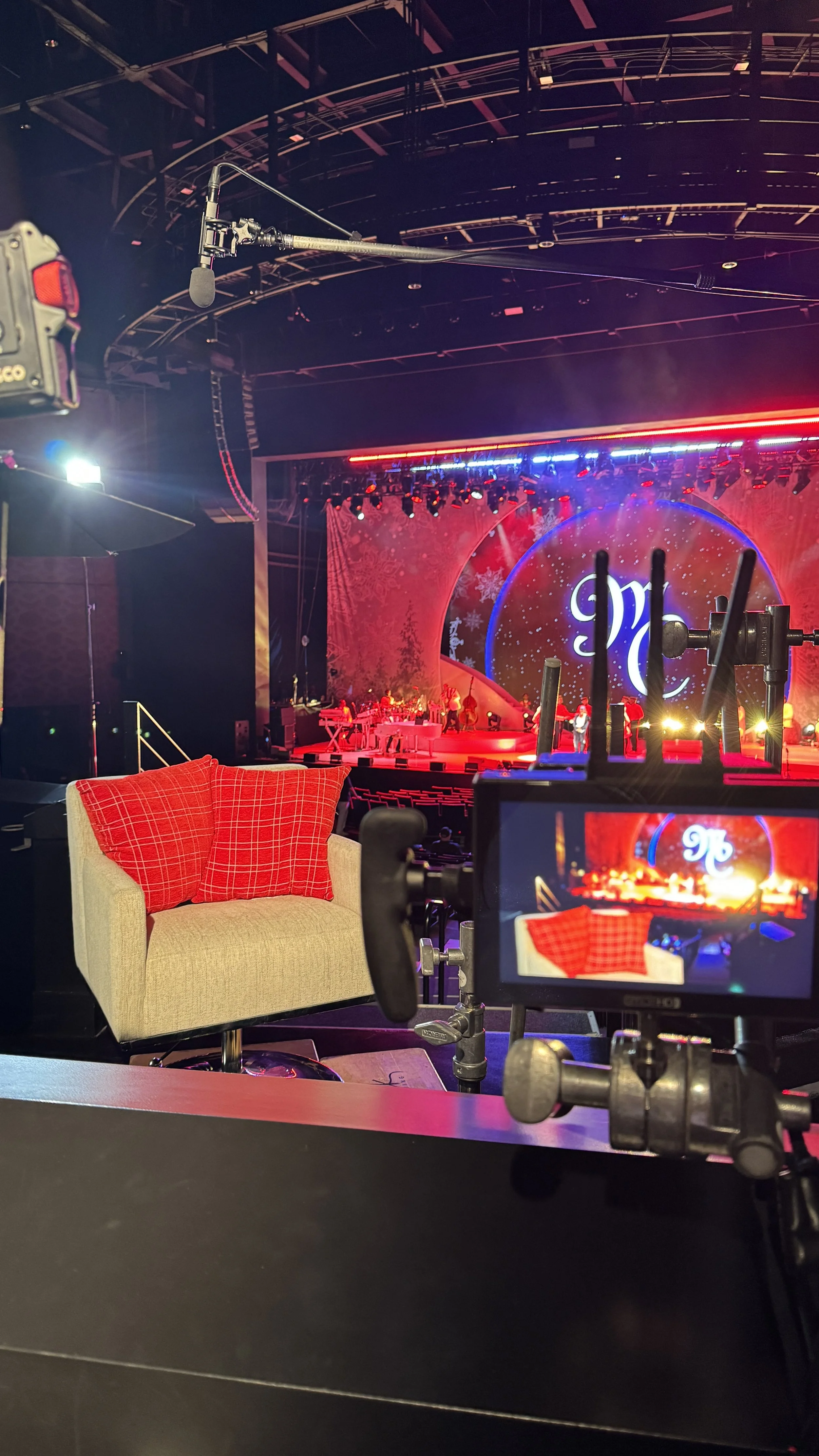 TV studio set with armchair and camera setup in foreground, stage with Christmas-themed decorations and lighting in background.
