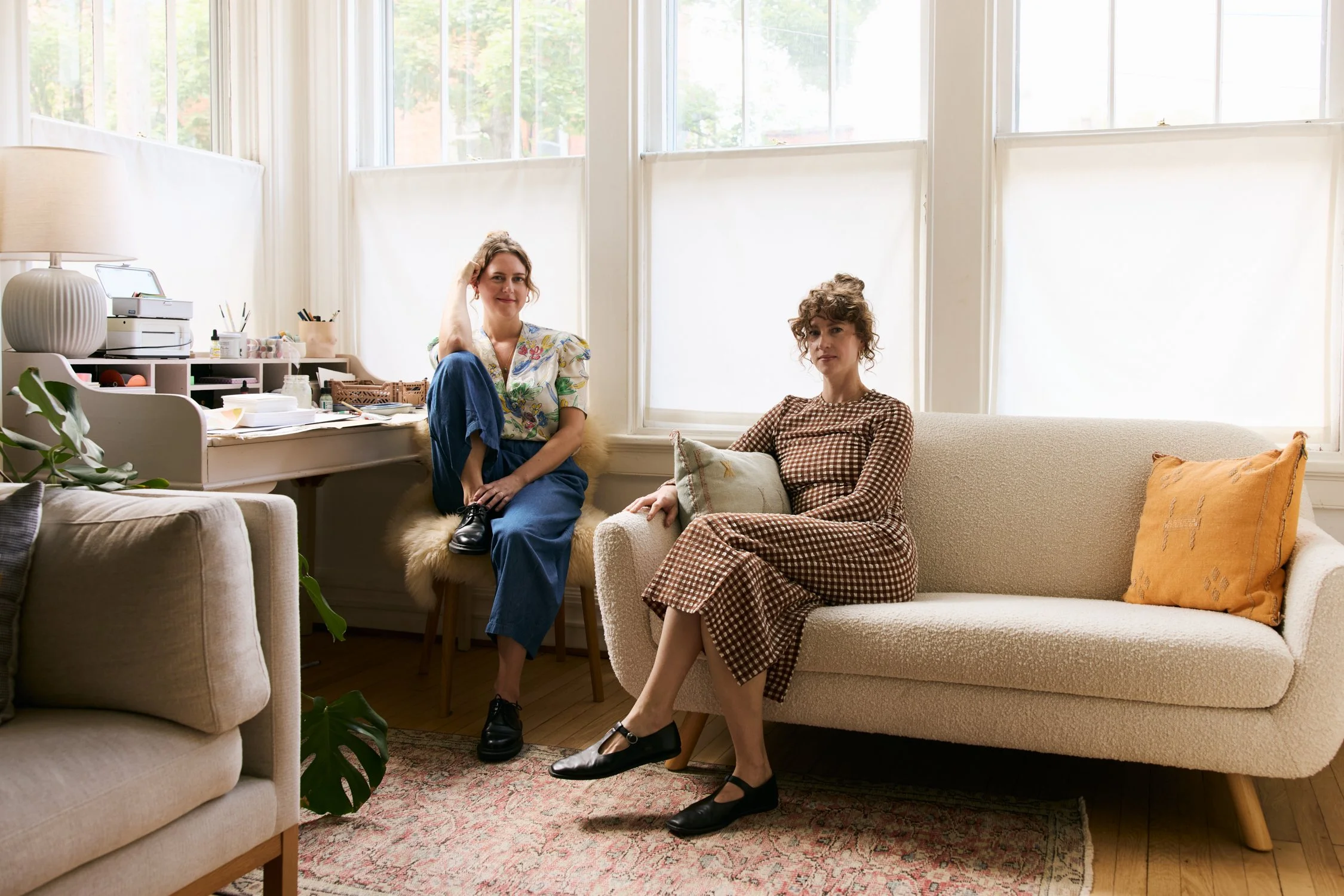 Two women are sitting inside a well-lit living room with large windows. One woman is sitting on a beige couch, wearing a brown checkered dress and black shoes, while the other woman is sitting on a chair or small sofa, wearing a floral top and blue pants. The room has a cream-colored sofa, a patterned rug, and various decorative items.